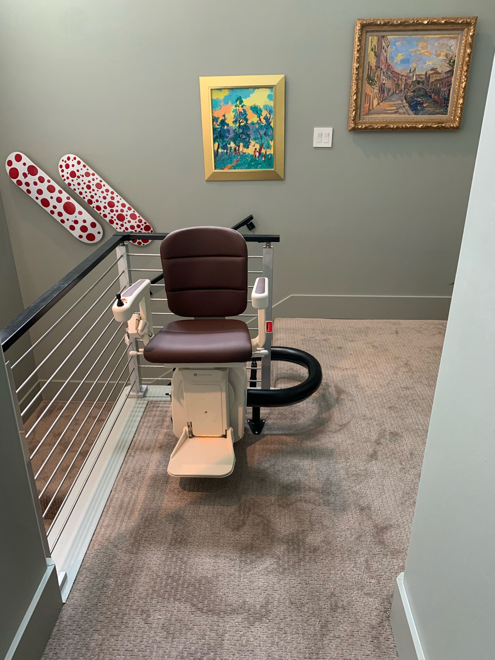 A brown chair stairlift at the top of a carpeted staircase in a room with framed art and wall-mounted red-dotted boards.