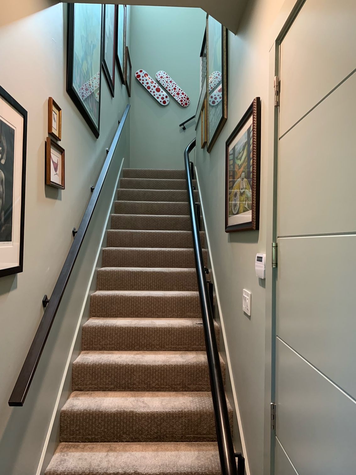 A staircase with carpeted steps, dark handrails, and framed artwork on light green walls leading to a bright landing.