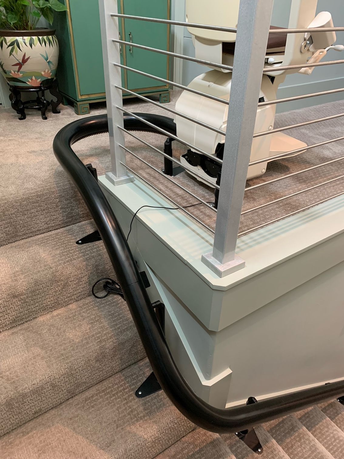 A stairlift track curves along carpeted stairs near a landing with a cable railing and a decorative plant.