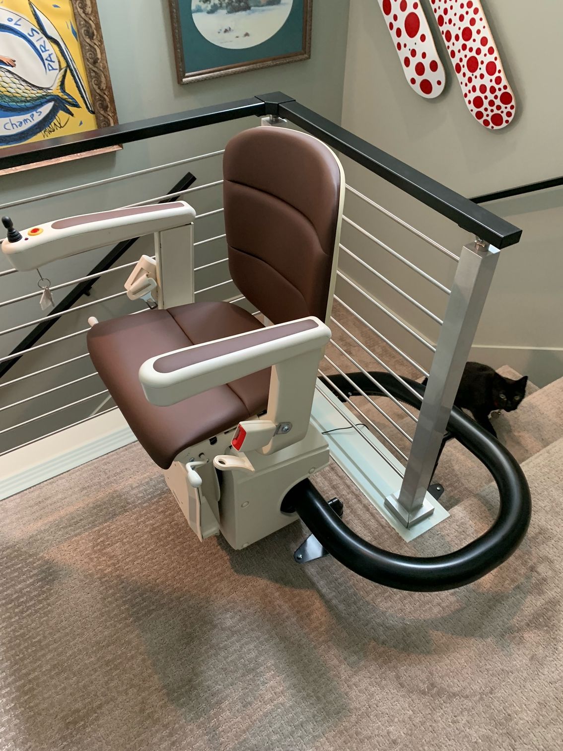 A brown and beige stair lift positioned at the top of a carpeted staircase next to a black cat and a railing.