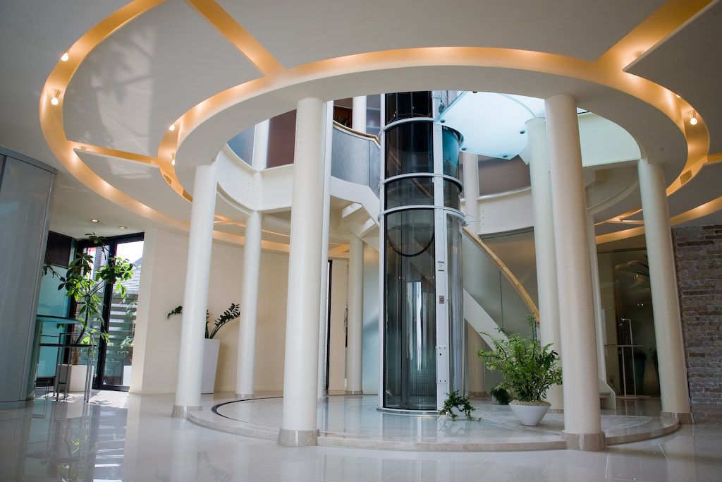 A bright, modern circular foyer featuring white columns, marble flooring, and a central glass cylindrical elevator.