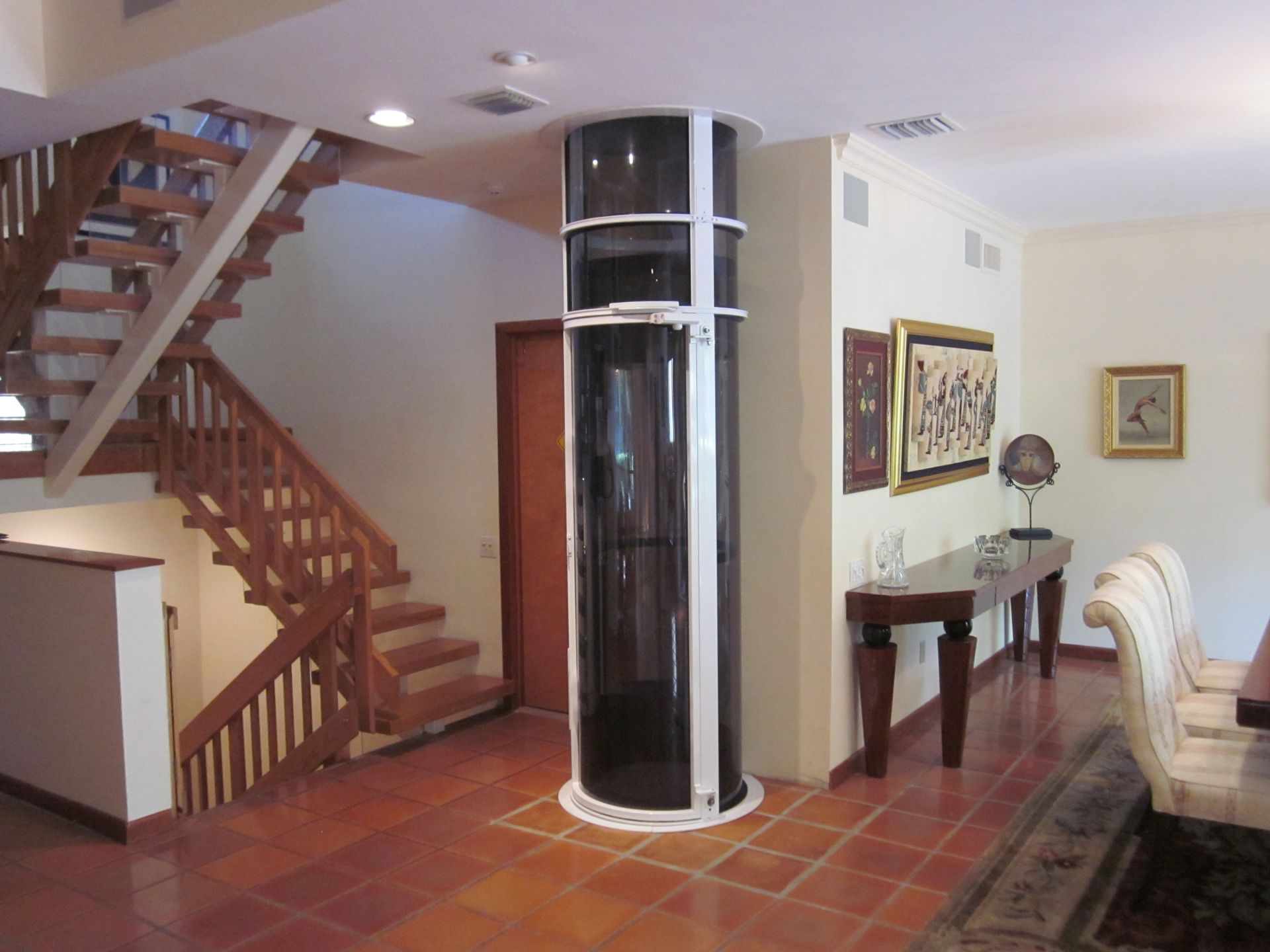A white cylindrical glass elevator stands in a foyer near a wooden staircase, a dining table, and terracotta tile floors.