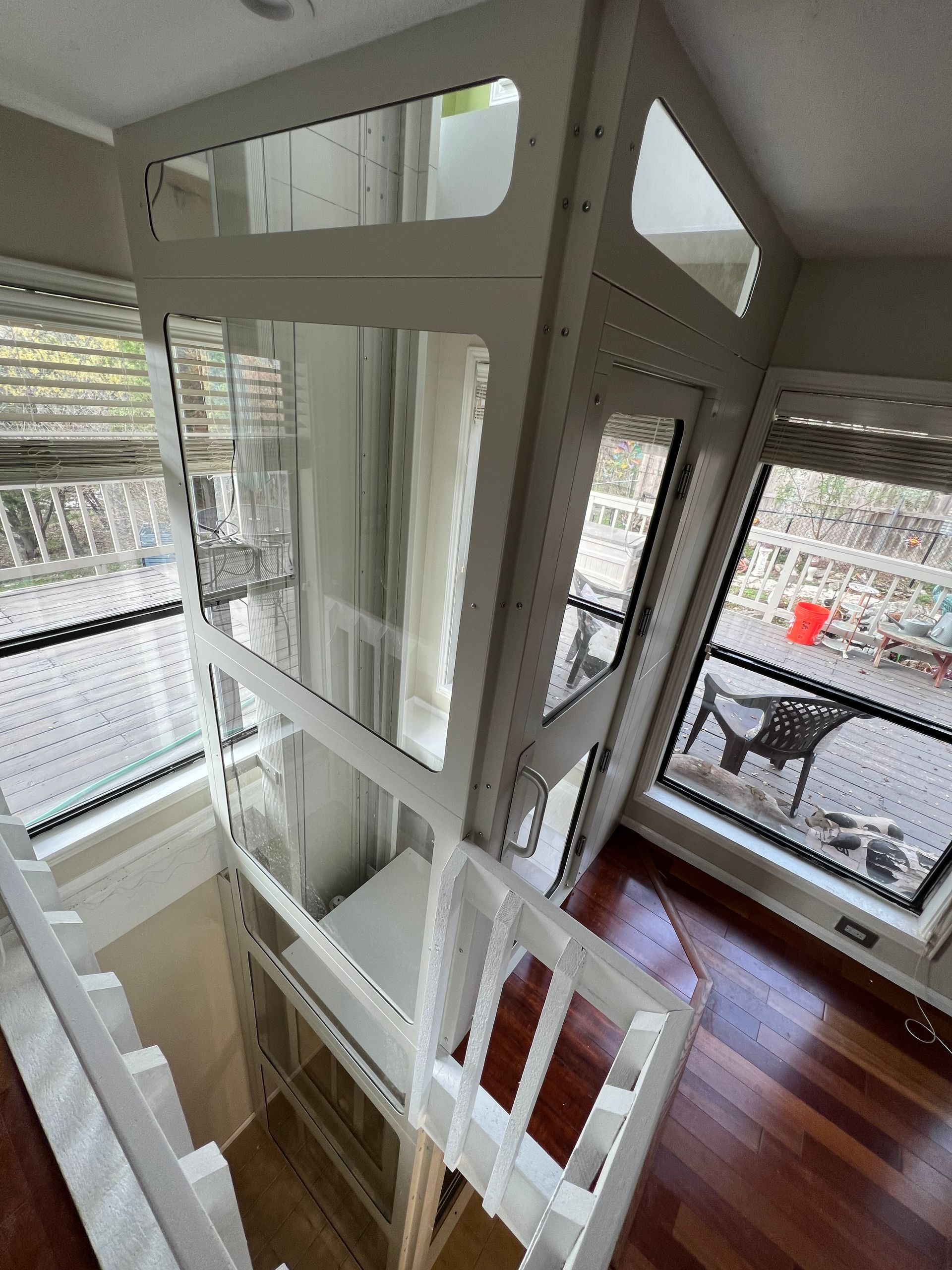 A white residential elevator with glass panels installed in a room with hardwood floors and large windows.