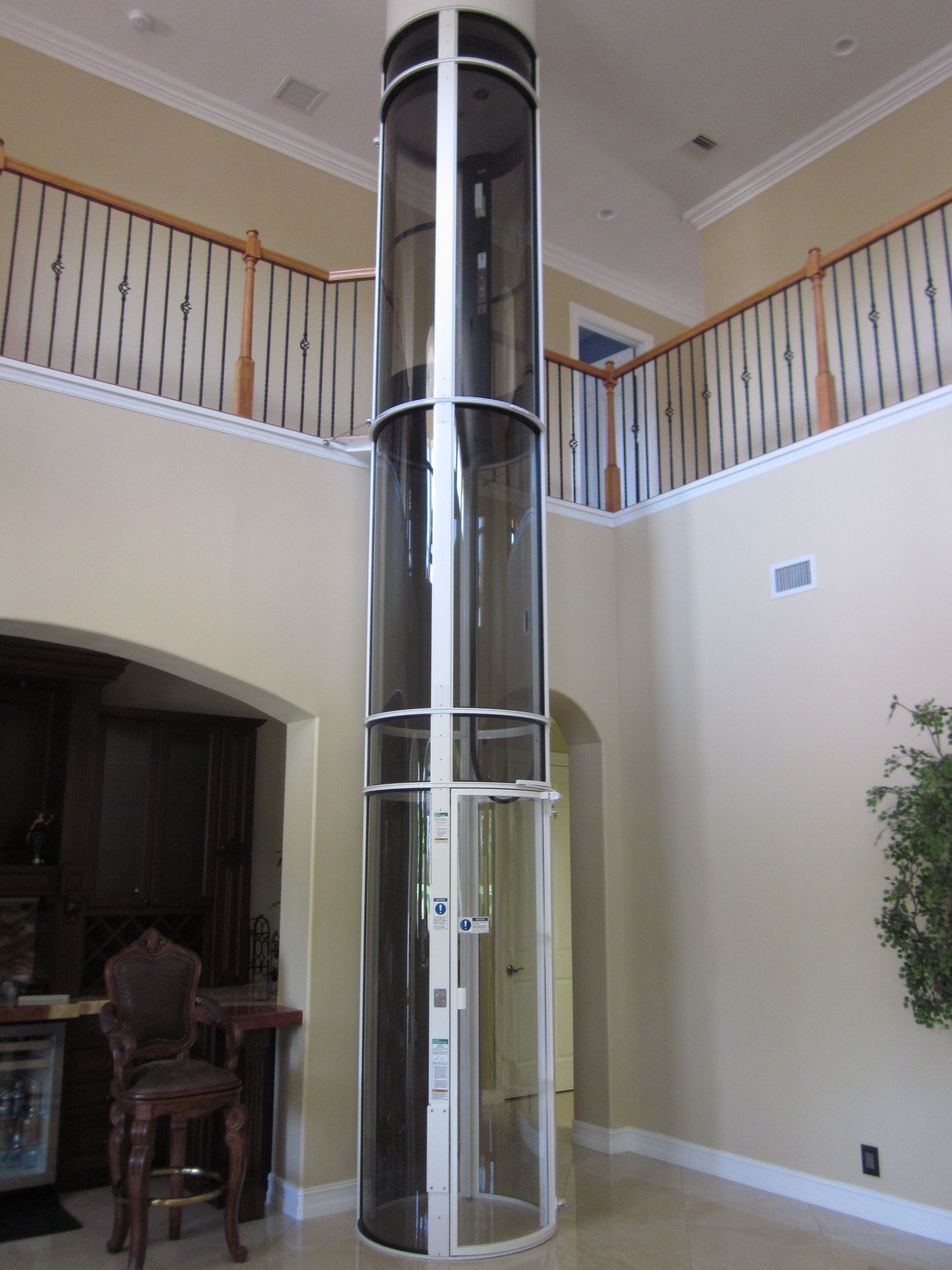 A cylindrical, glass-walled residential elevator stands in the center of an open, two-story home foyer.