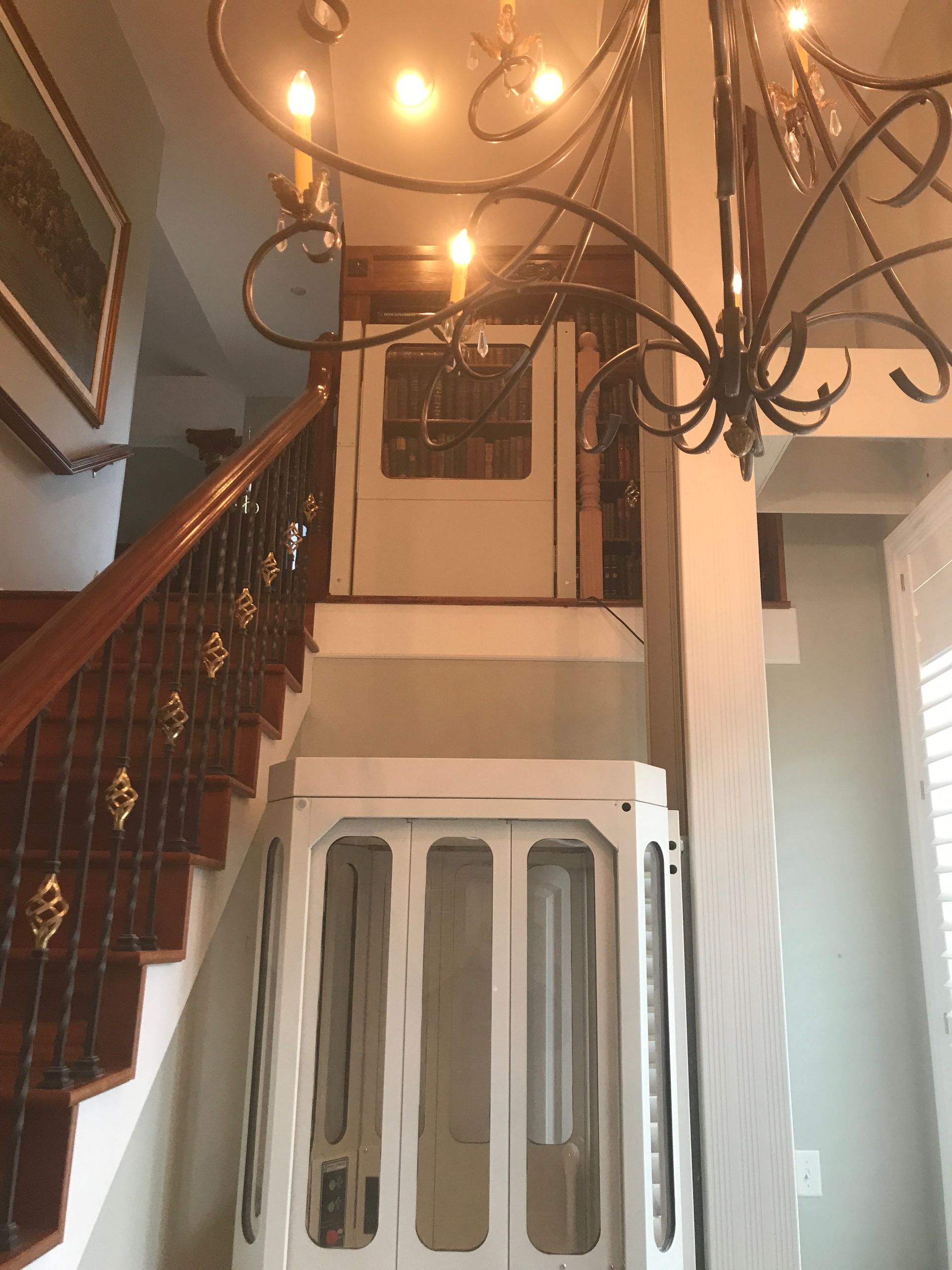 A grand chandelier hangs above a home entryway with a wooden staircase and a white, glass-paned cabinet.