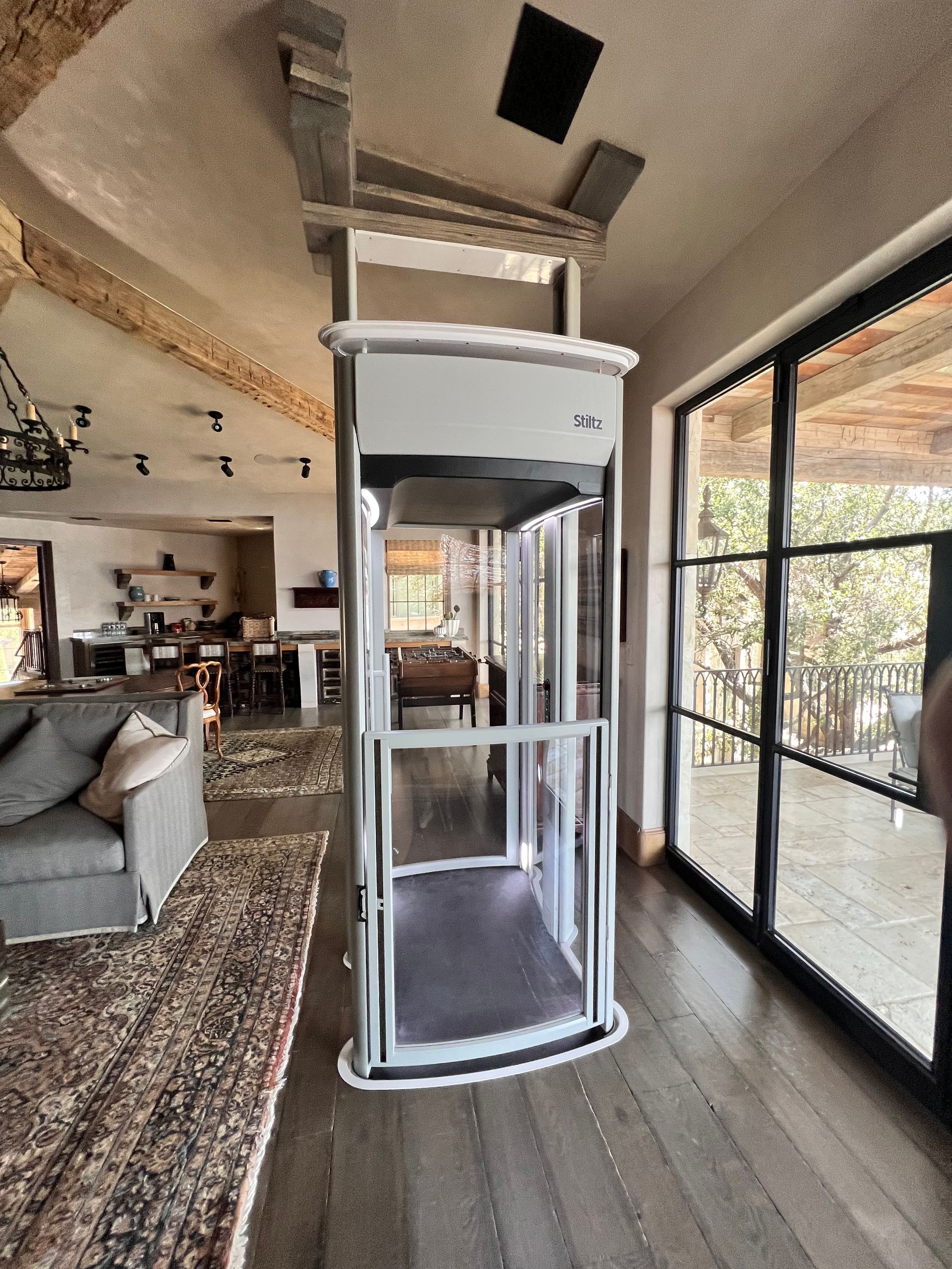 A residential hallway with wooden doors and floors, featuring a modern, cylindrical glass home elevator as a central focus.