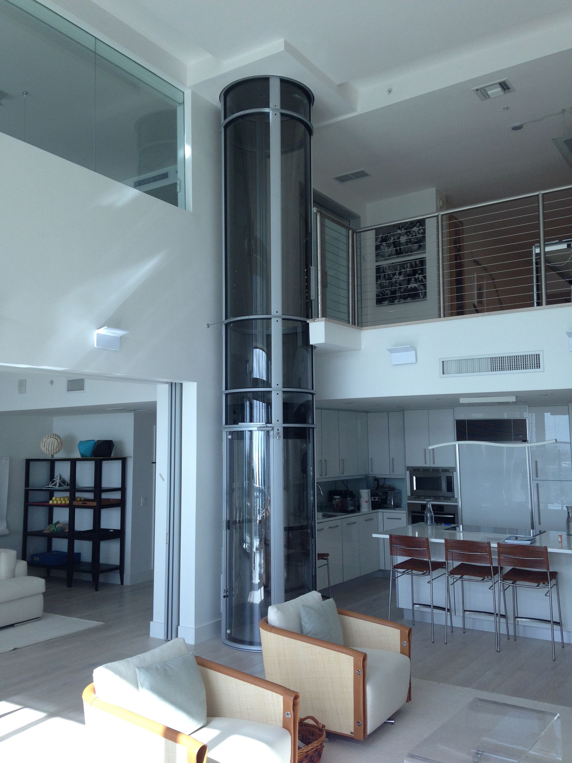A modern, cylindrical glass home elevator stands in the center of an open-concept living area with white decor.