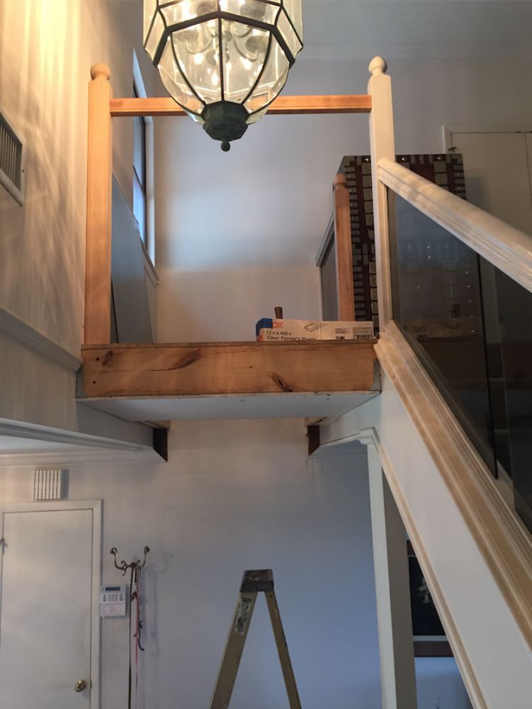 A view of a stairwell landing under renovation, featuring a raw wooden beam, railing posts, and an overhead pendant light.