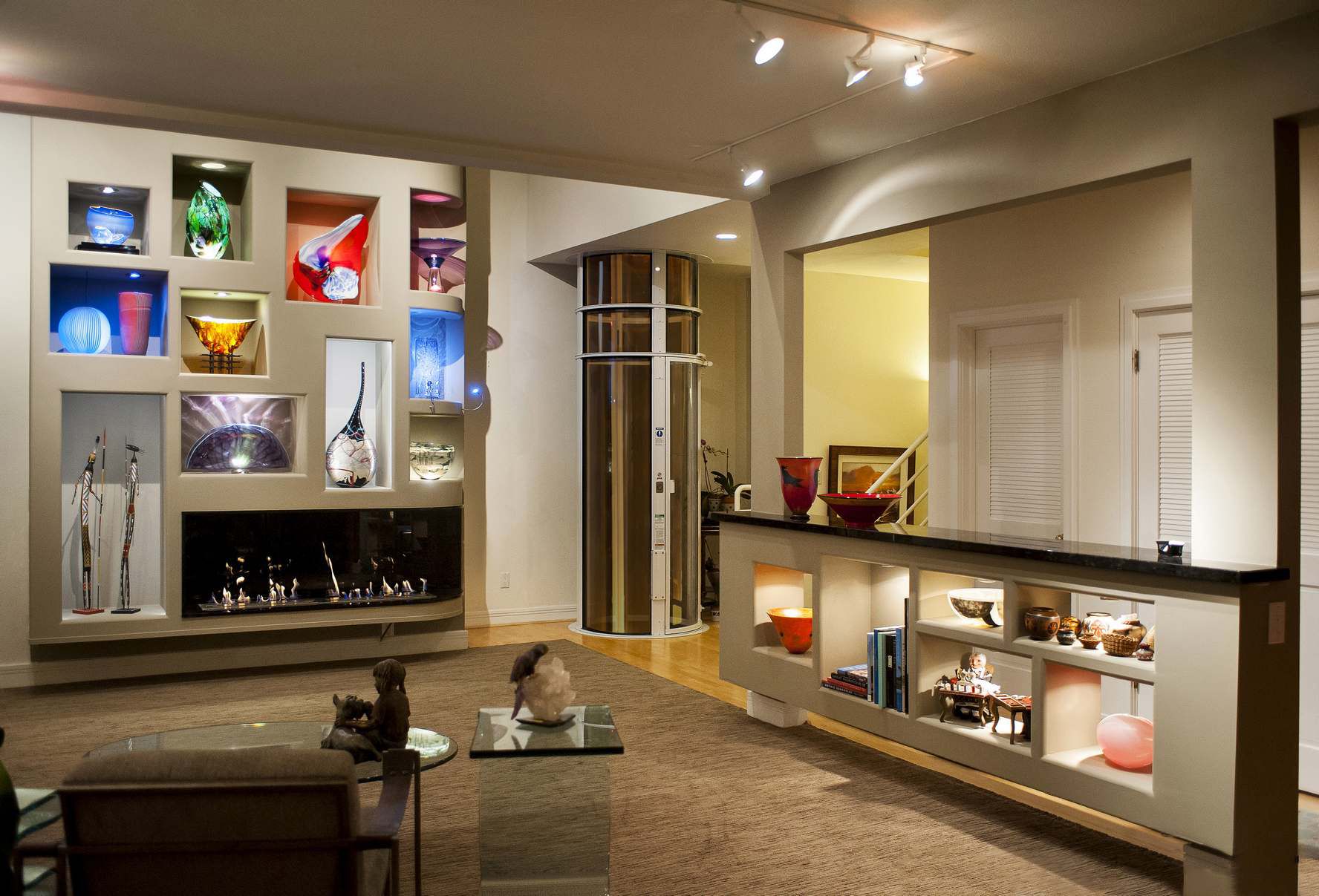 A modern living room with a large, illuminated shelving unit for art, a linear fireplace, and a glass-enclosed elevator.