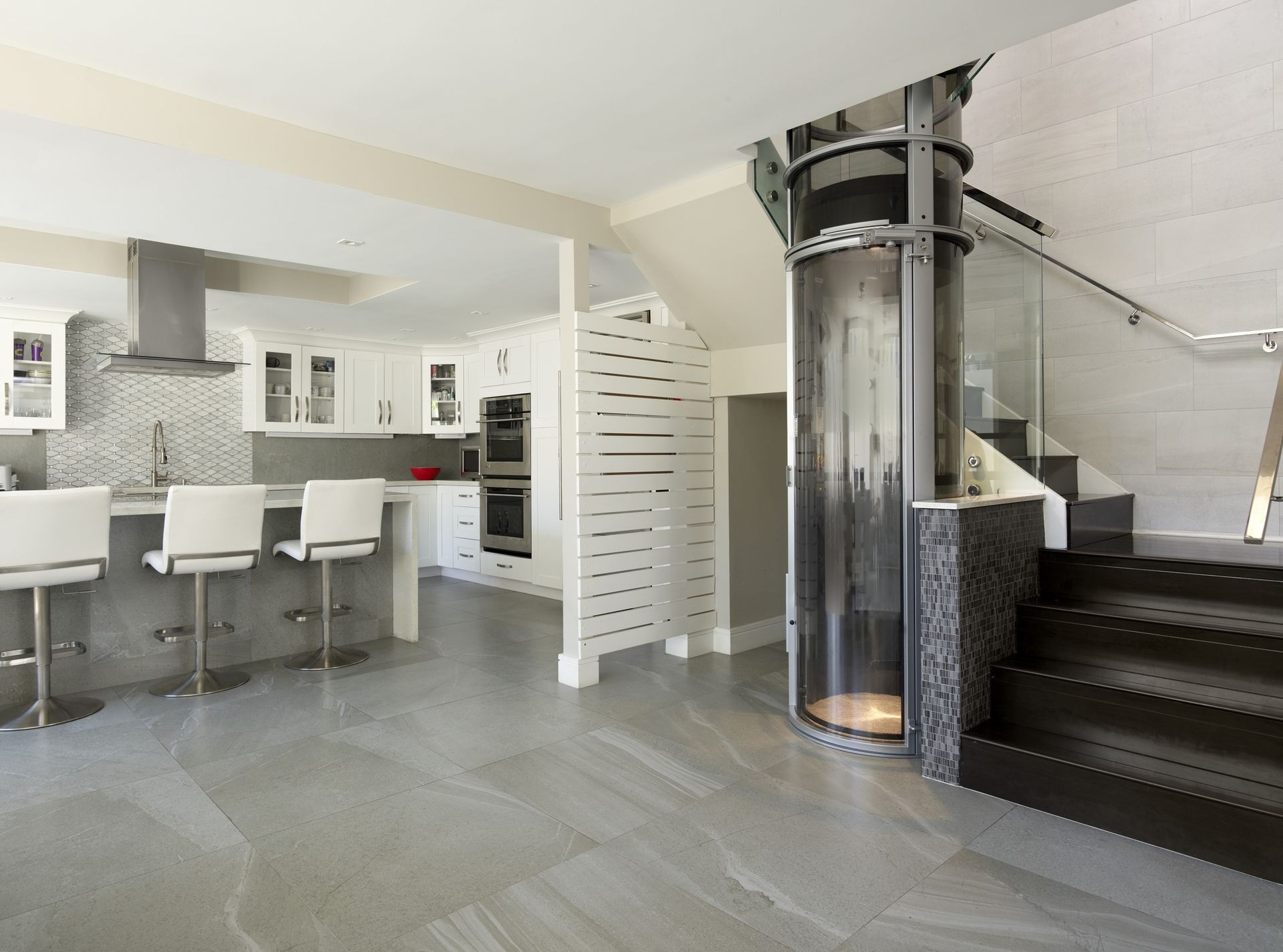 Modern kitchen with white cabinets and a breakfast bar, featuring a circular glass elevator next to a wooden staircase.