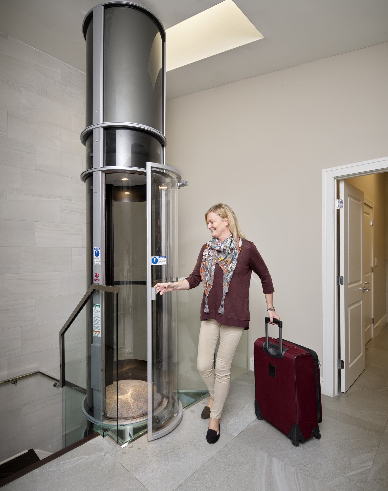 A person stands by an open cylindrical home elevator, holding a large red suitcase in a modern, bright indoor setting.