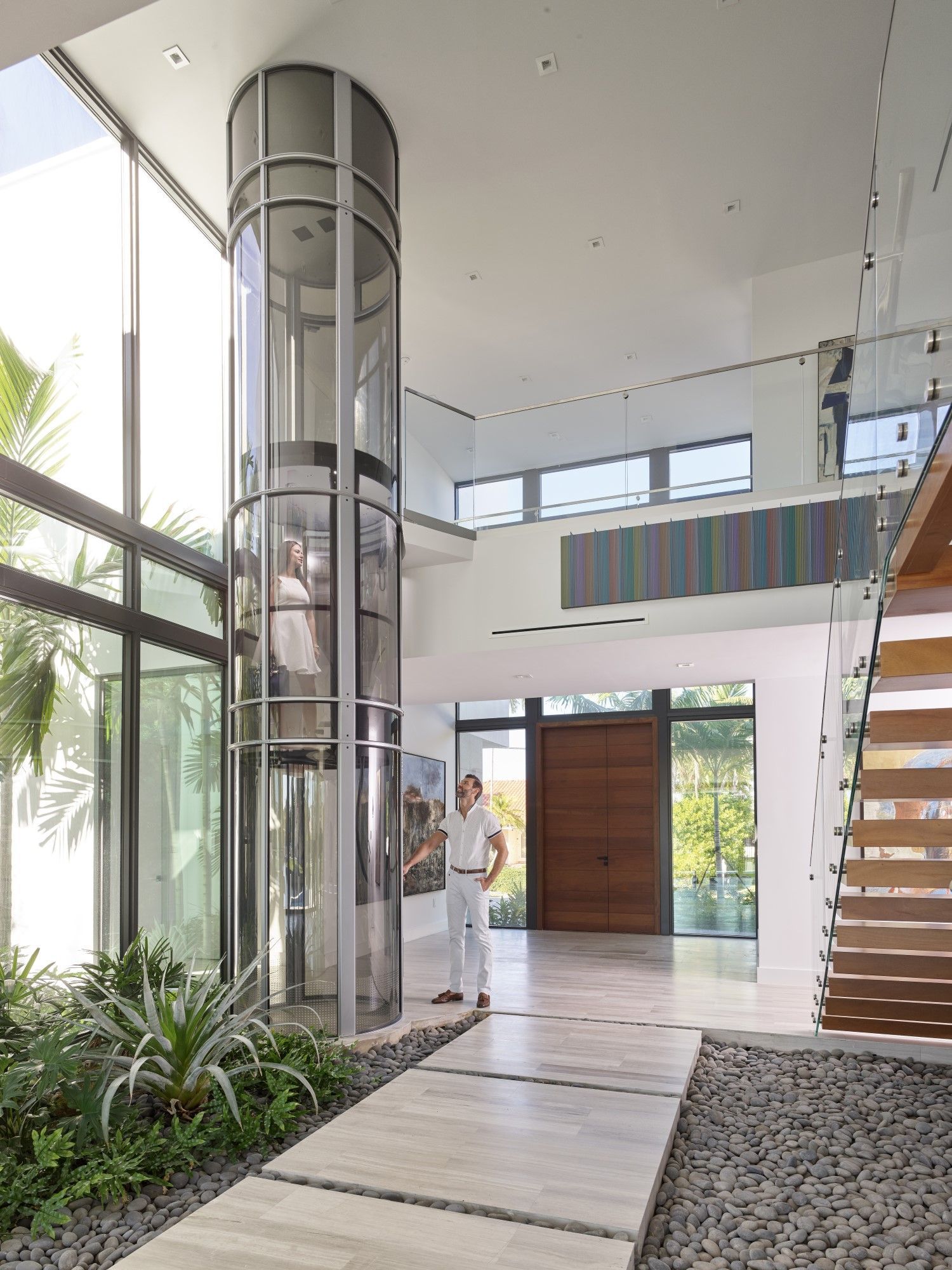 A person stands in a bright, modern foyer next to a tall, cylindrical glass elevator.
