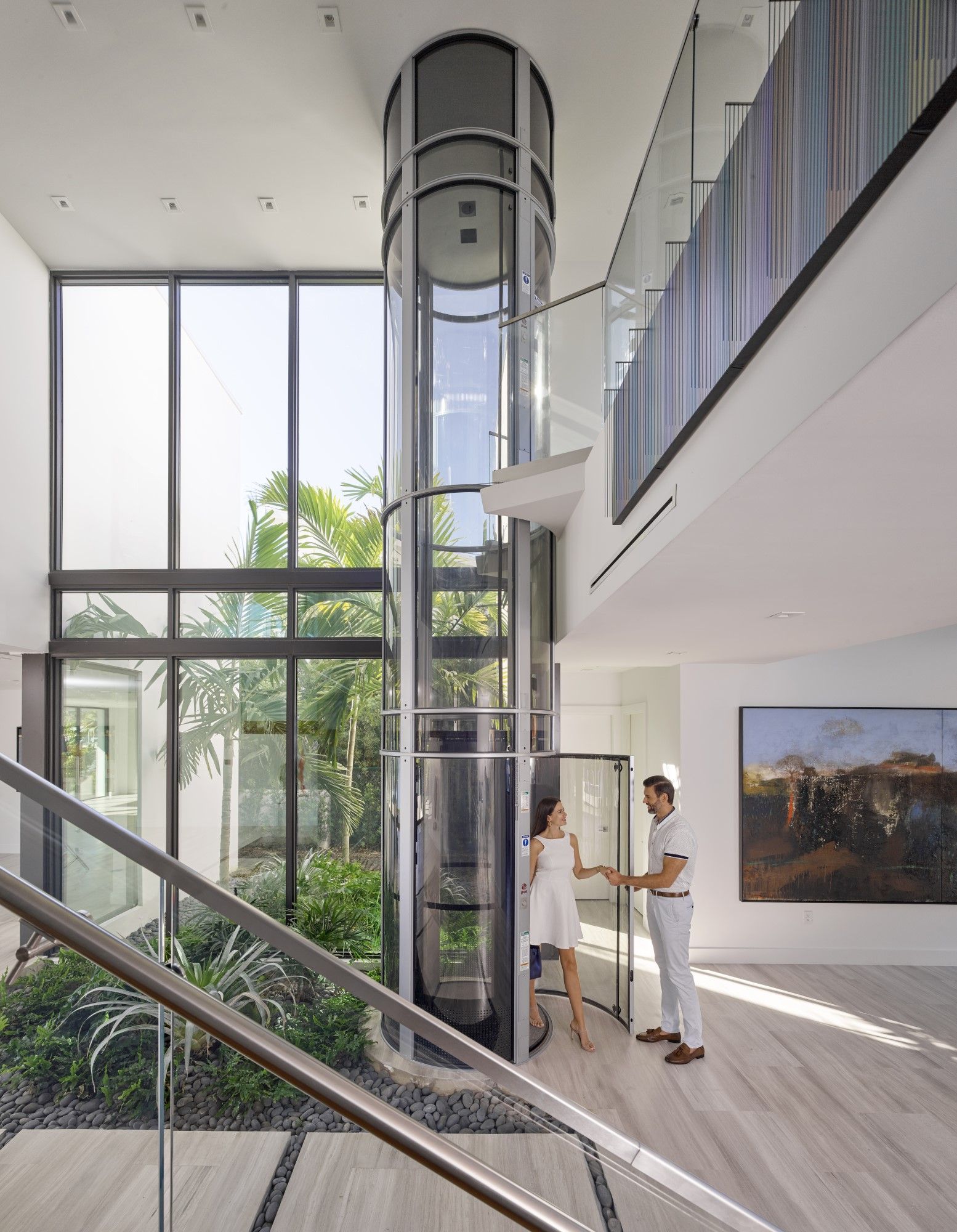A modern home interior features a central glass cylindrical elevator with a person standing by its open door.