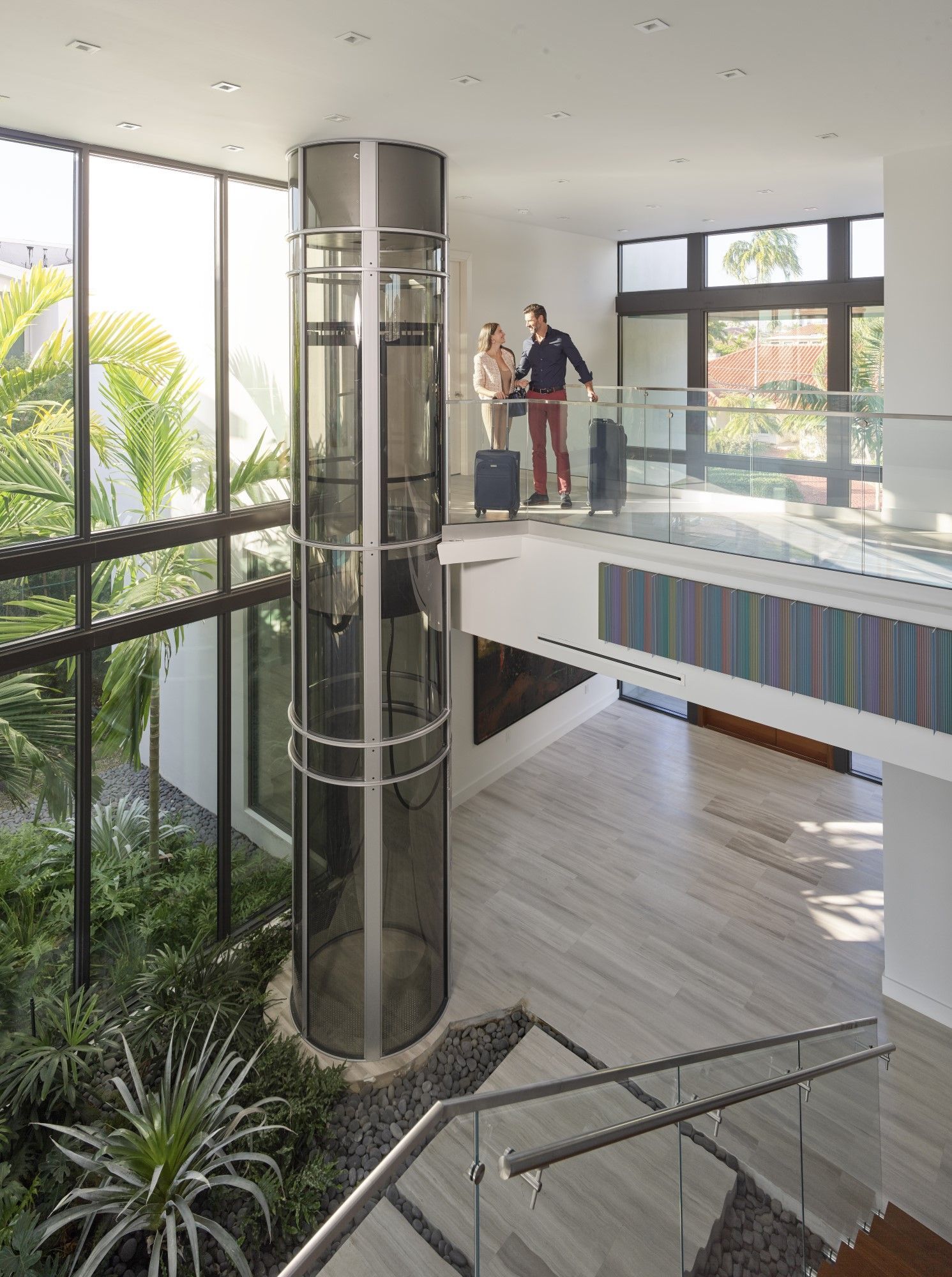 Two people with luggage stand on a modern, open indoor balcony next to a cylindrical glass elevator in a sunny atrium.