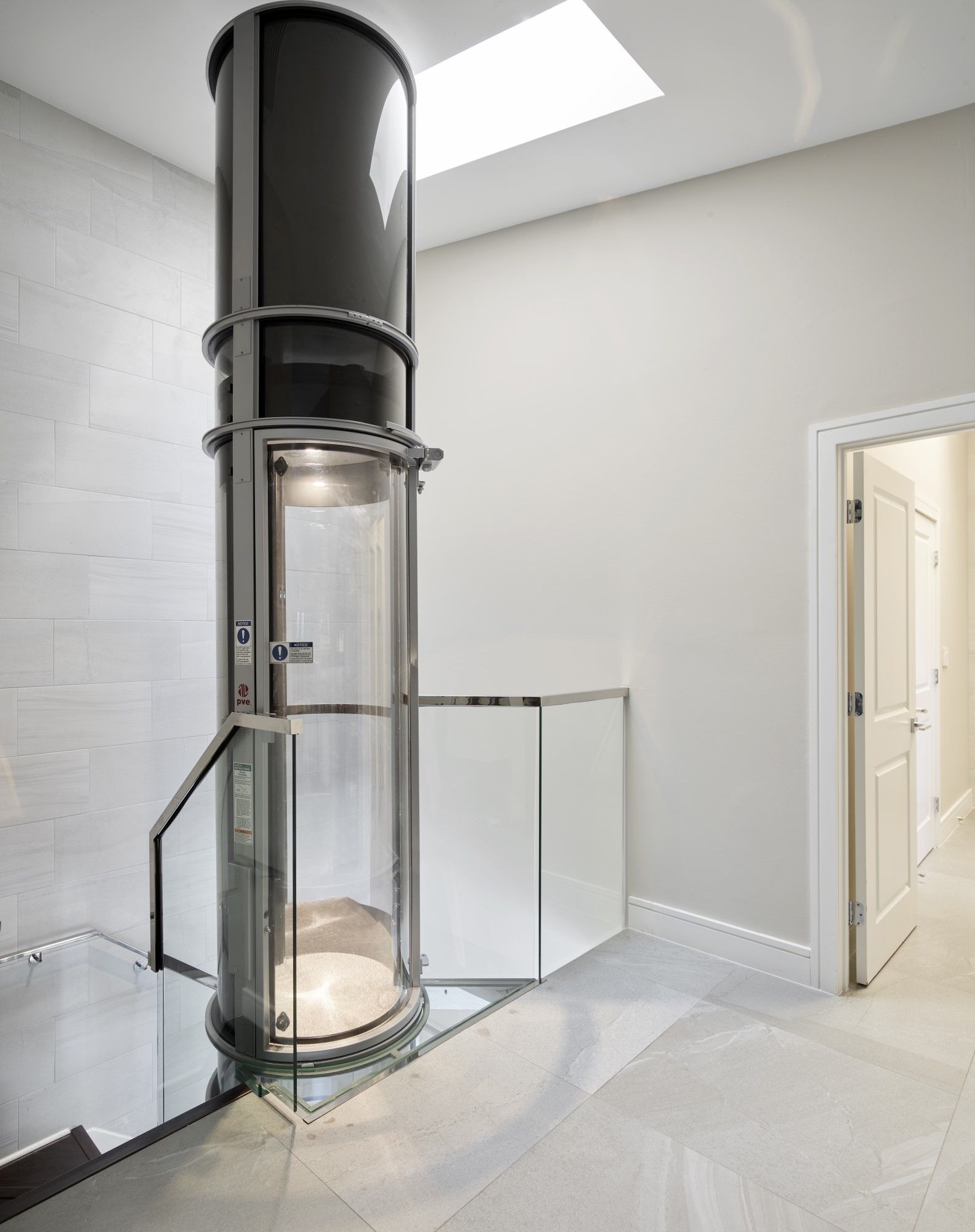 A modern, cylindrical glass home elevator installed in a minimalist room with white walls, tile floors, and a skylight.