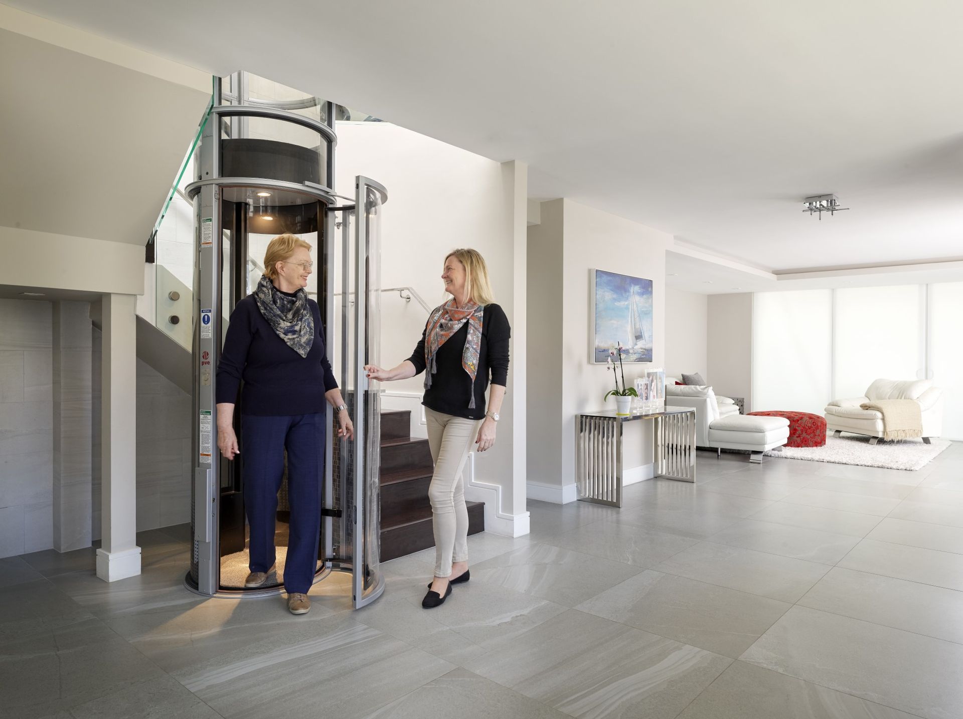 Two people stand near a glass, cylindrical home elevator inside a modern, neutrally-colored living space.