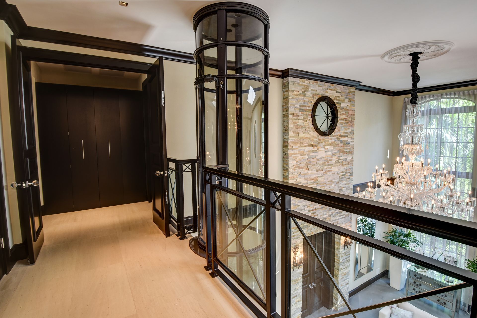 A modern indoor glass elevator stands in a hallway beside a black railing overlooking a room with a crystal chandelier.