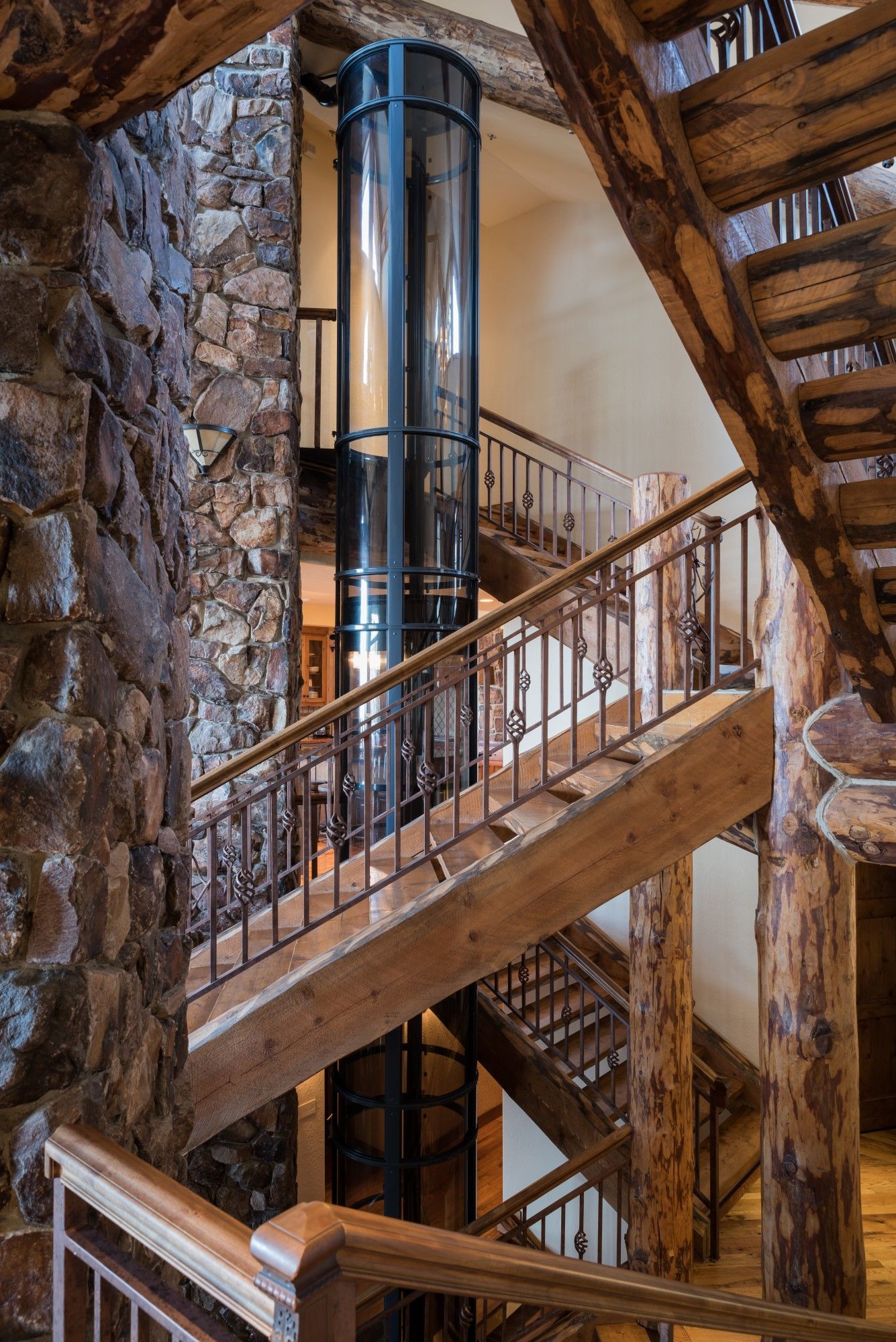 A cylindrical glass elevator stands in the center of a rustic wooden staircase and a tall stone wall in a home interior.