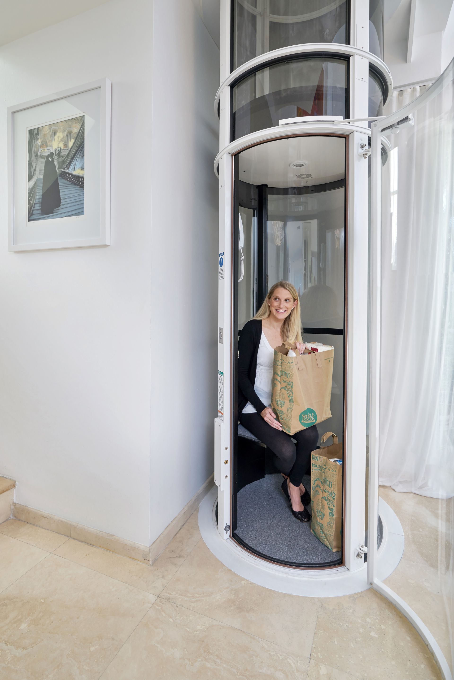 A woman with grocery bags sits inside a small, cylindrical glass home elevator installed in a bright, modern room.