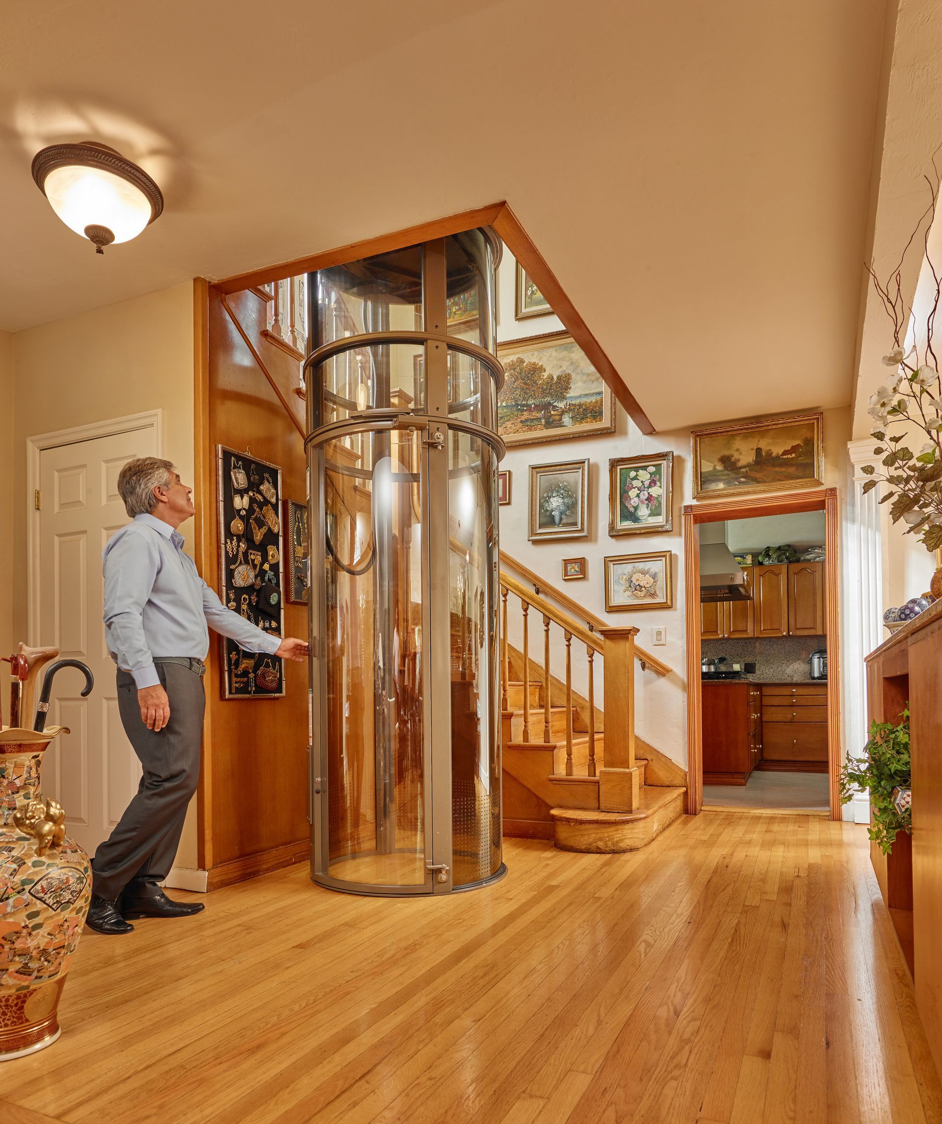 A person in a house standing next to a cylindrical, glass home elevator positioned near a wooden staircase.