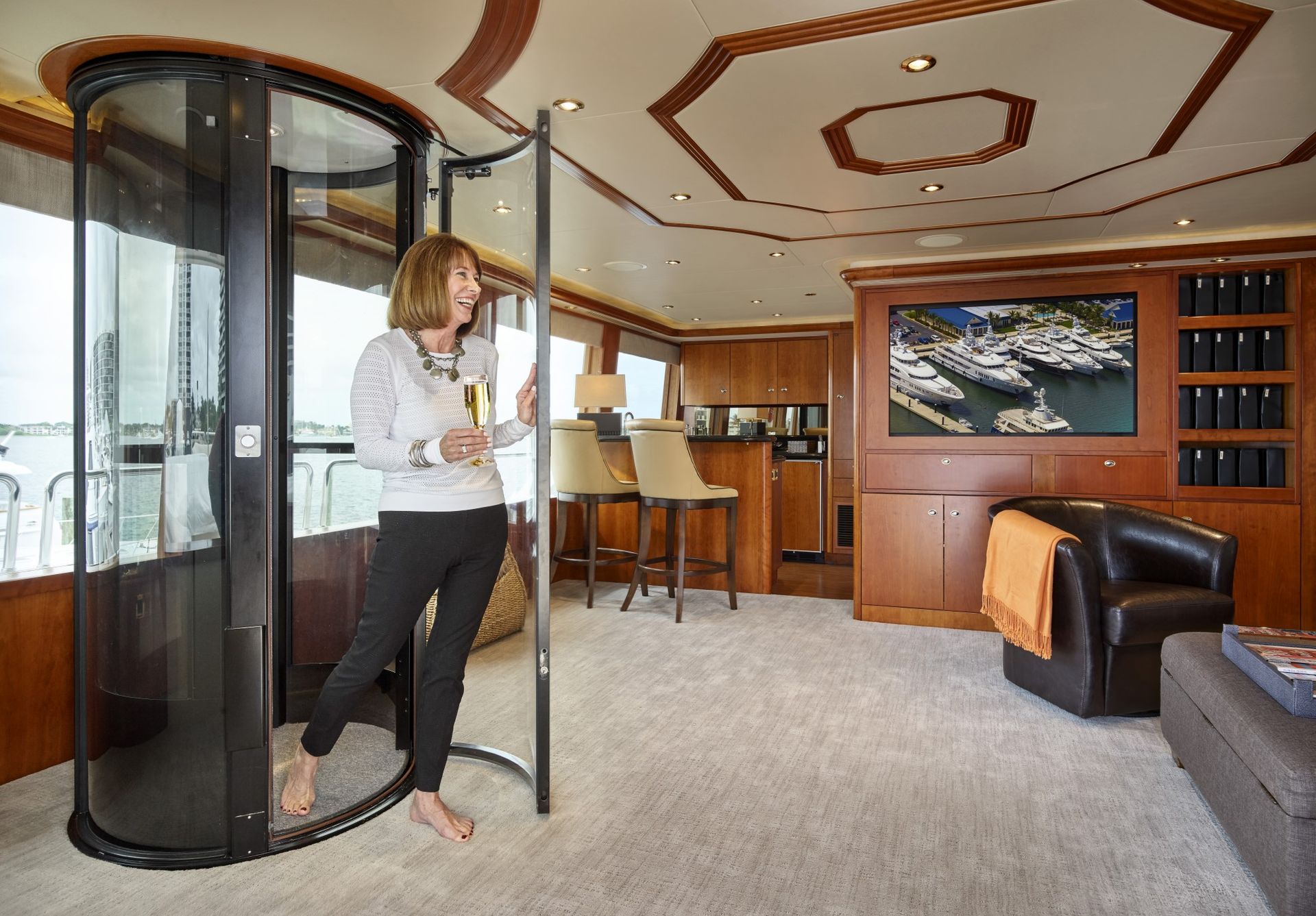 A person steps out of a glass cylindrical elevator into a wood-paneled interior room with furniture and a large TV.