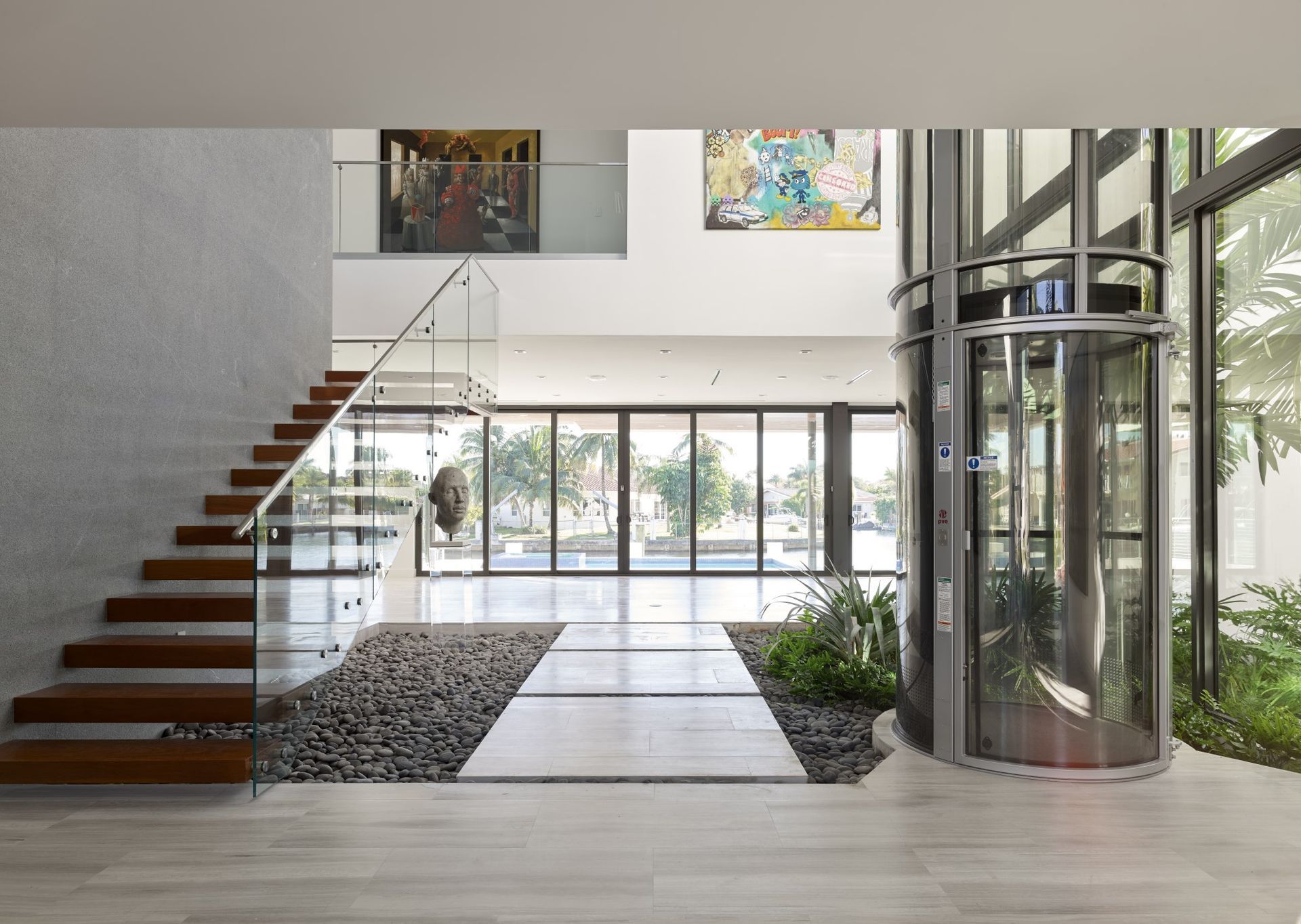 A modern foyer with wood-tread stairs, a glass cylindrical elevator, stone flooring, and a small indoor garden area.