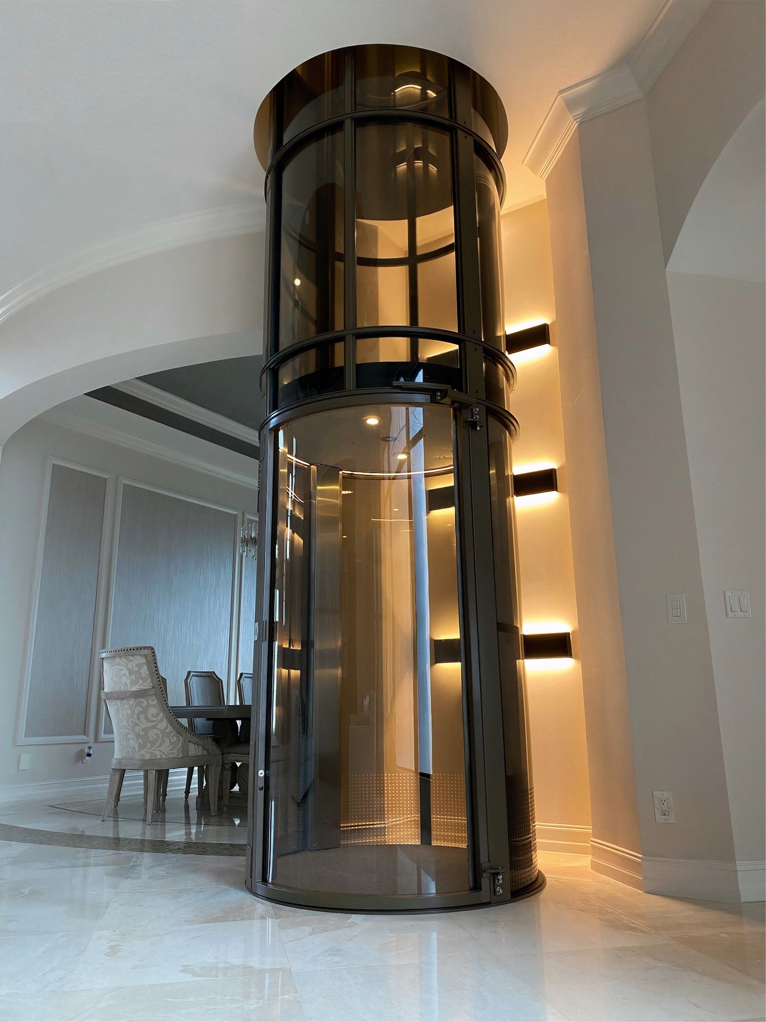 A modern, cylindrical glass home elevator with a dark metal frame stands in a brightly lit, elegant residential room.