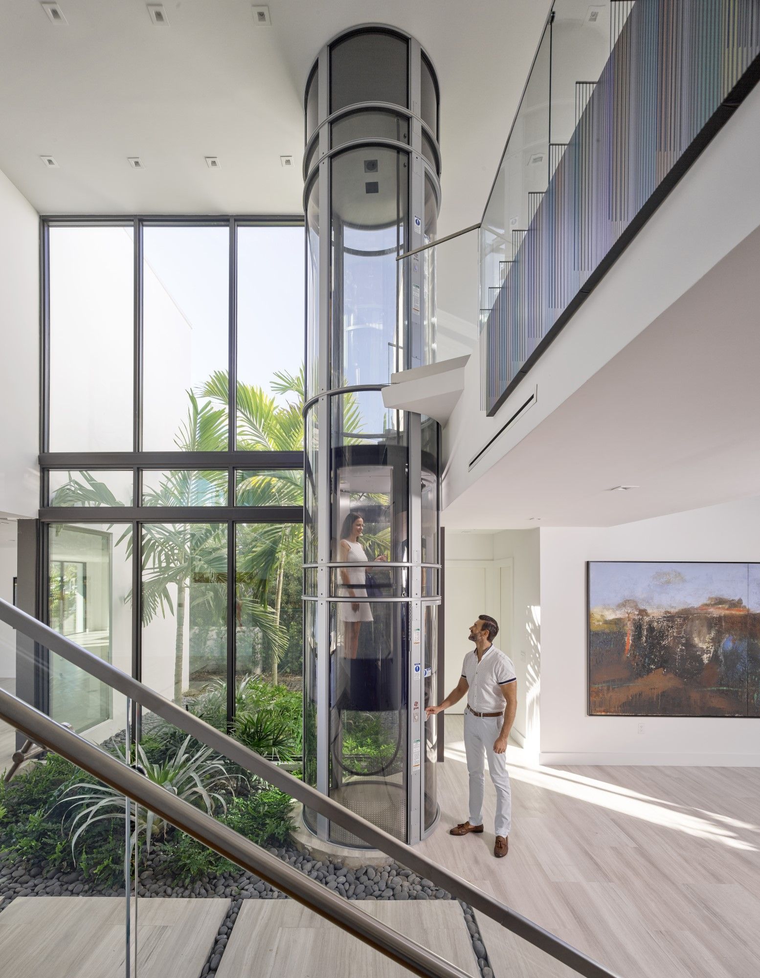 A person stands in a modern, bright home looking at a cylindrical glass elevator with a person inside.