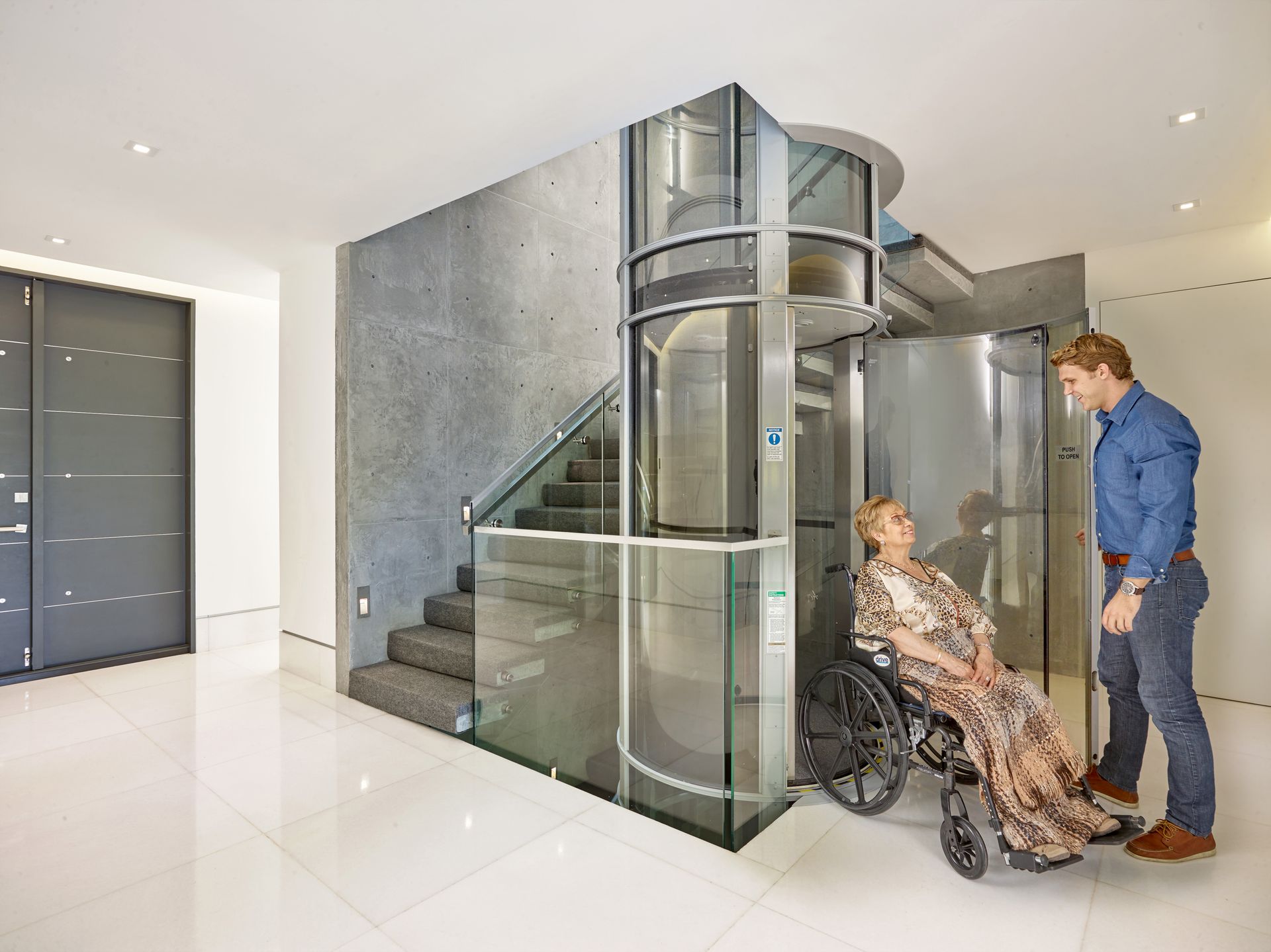 A person in a wheelchair assisted by another person at a modern, glass-enclosed residential elevator next to stairs.