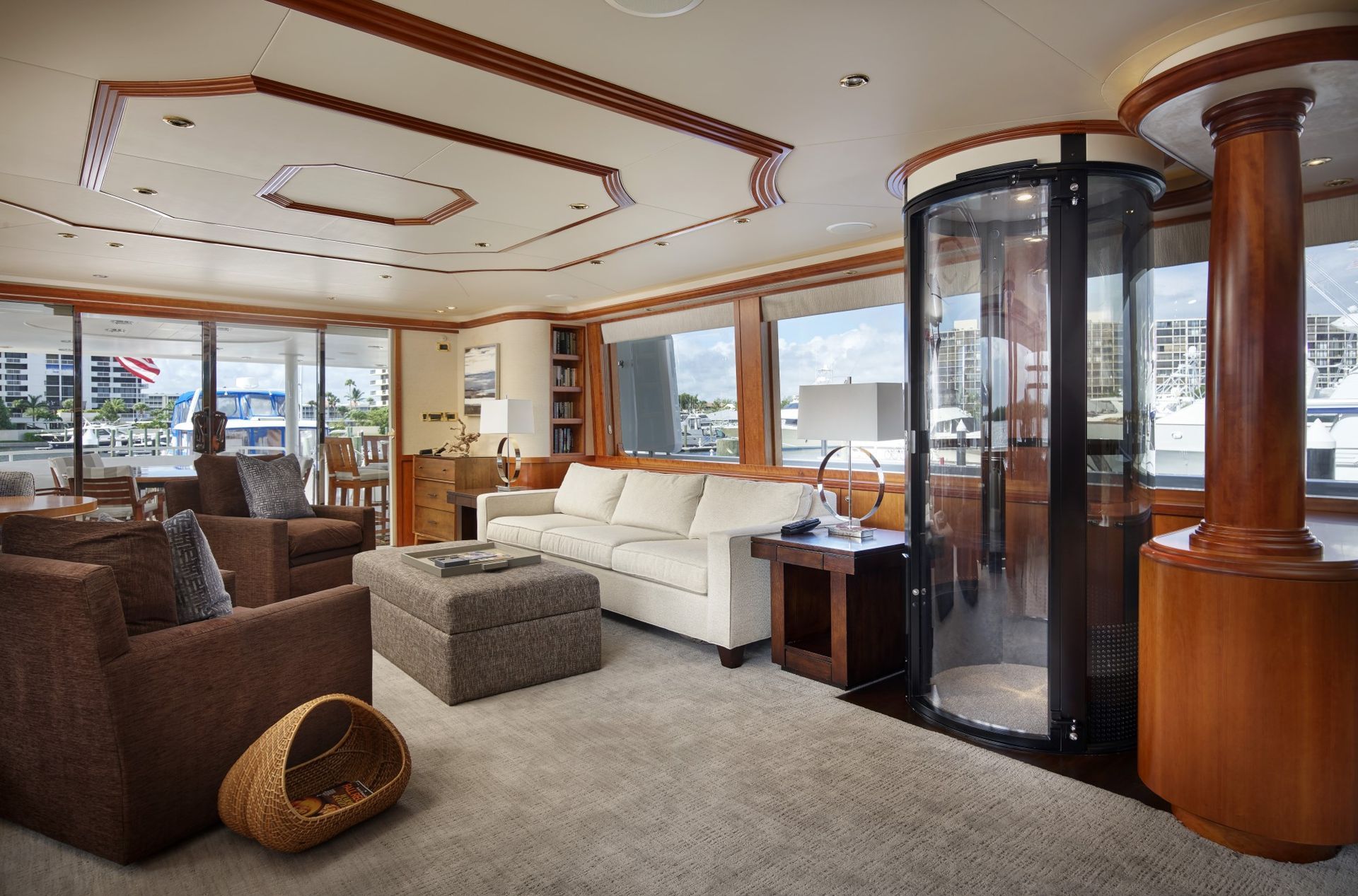 A luxurious yacht interior with a cream sofa, brown armchairs, a glass circular elevator, and wood trim near large windows.