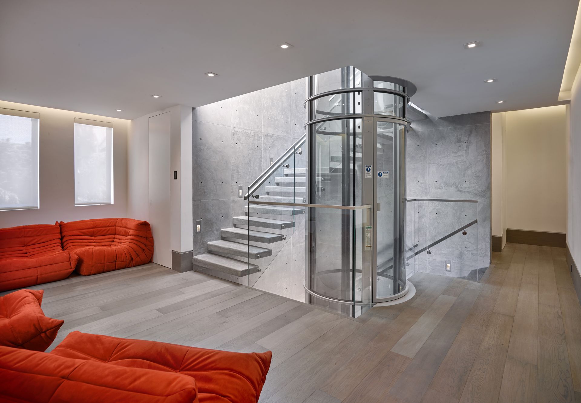 Modern interior with red modular seating, a glass cylindrical elevator, and concrete stairs against a concrete wall.