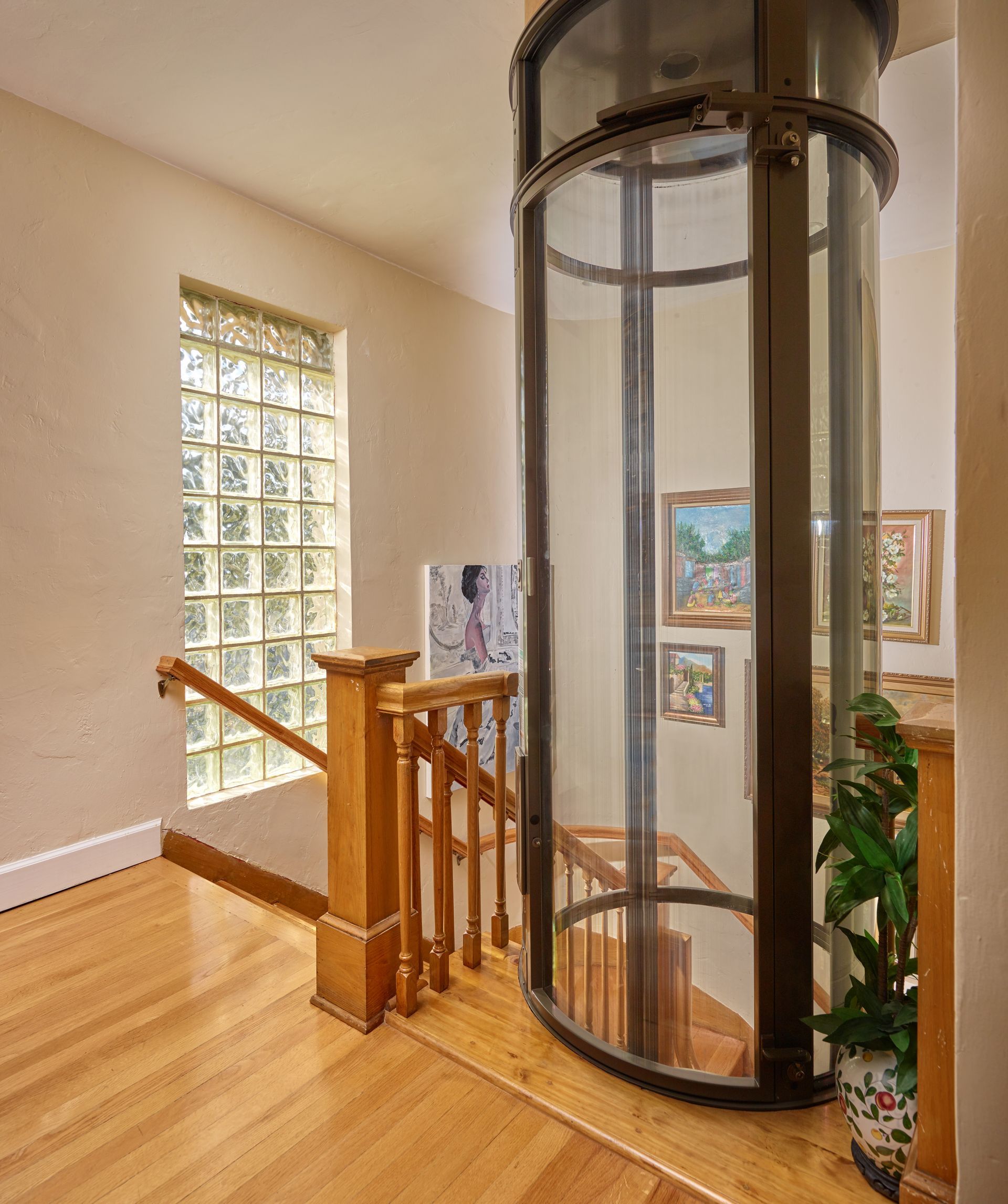 A cylindrical glass residential elevator stands beside a wooden staircase and glass block window in a home.