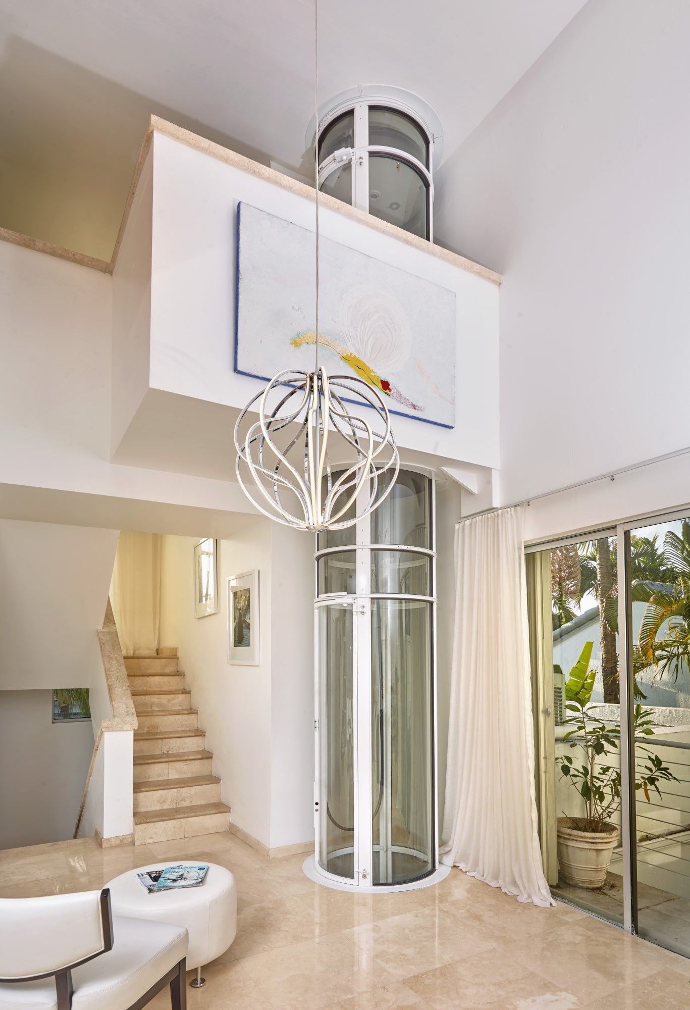 A modern, white, circular residential glass elevator stands in a bright, contemporary living space near a staircase.