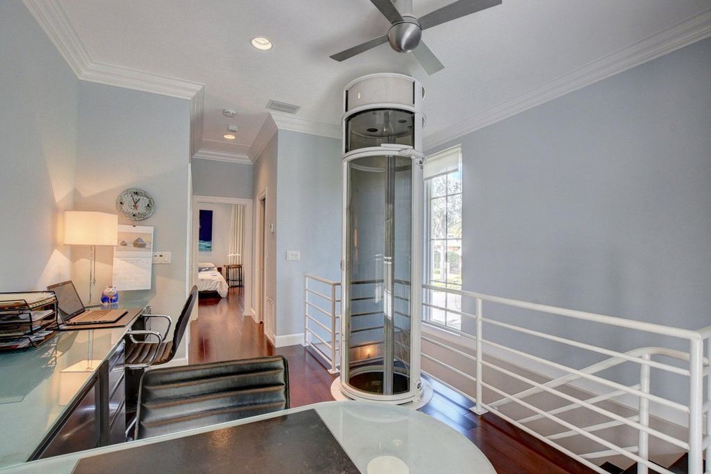 A home office with a glass cylindrical elevator, a desk, a ceiling fan, and a view into an adjacent bedroom.