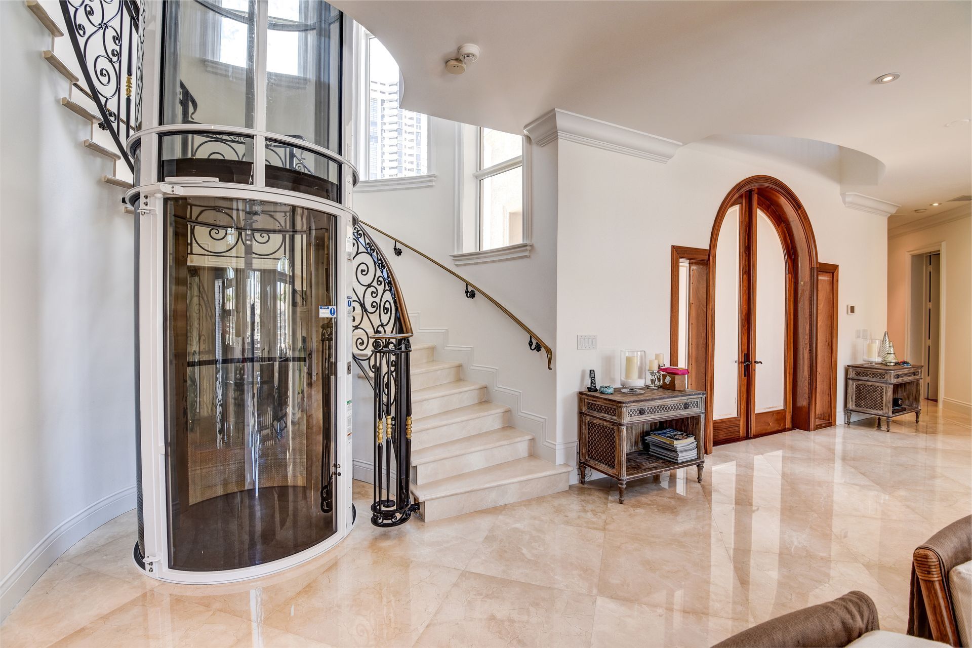 An ornate circular home elevator stands next to a curved marble staircase in a bright, elegant foyer with marble floors.