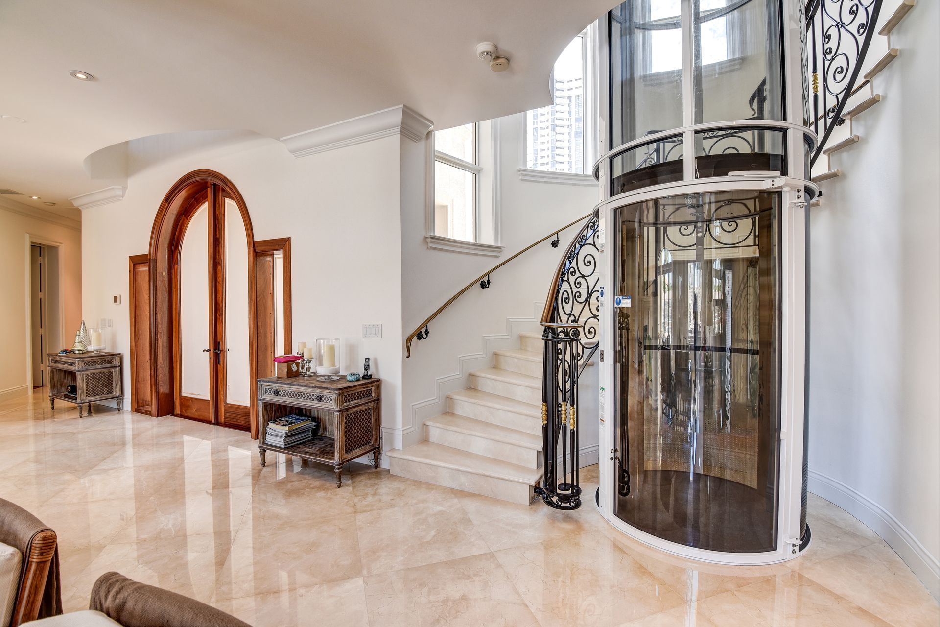 A spacious foyer with light tile floors, a white staircase with iron railings, and a cylindrical glass home elevator.