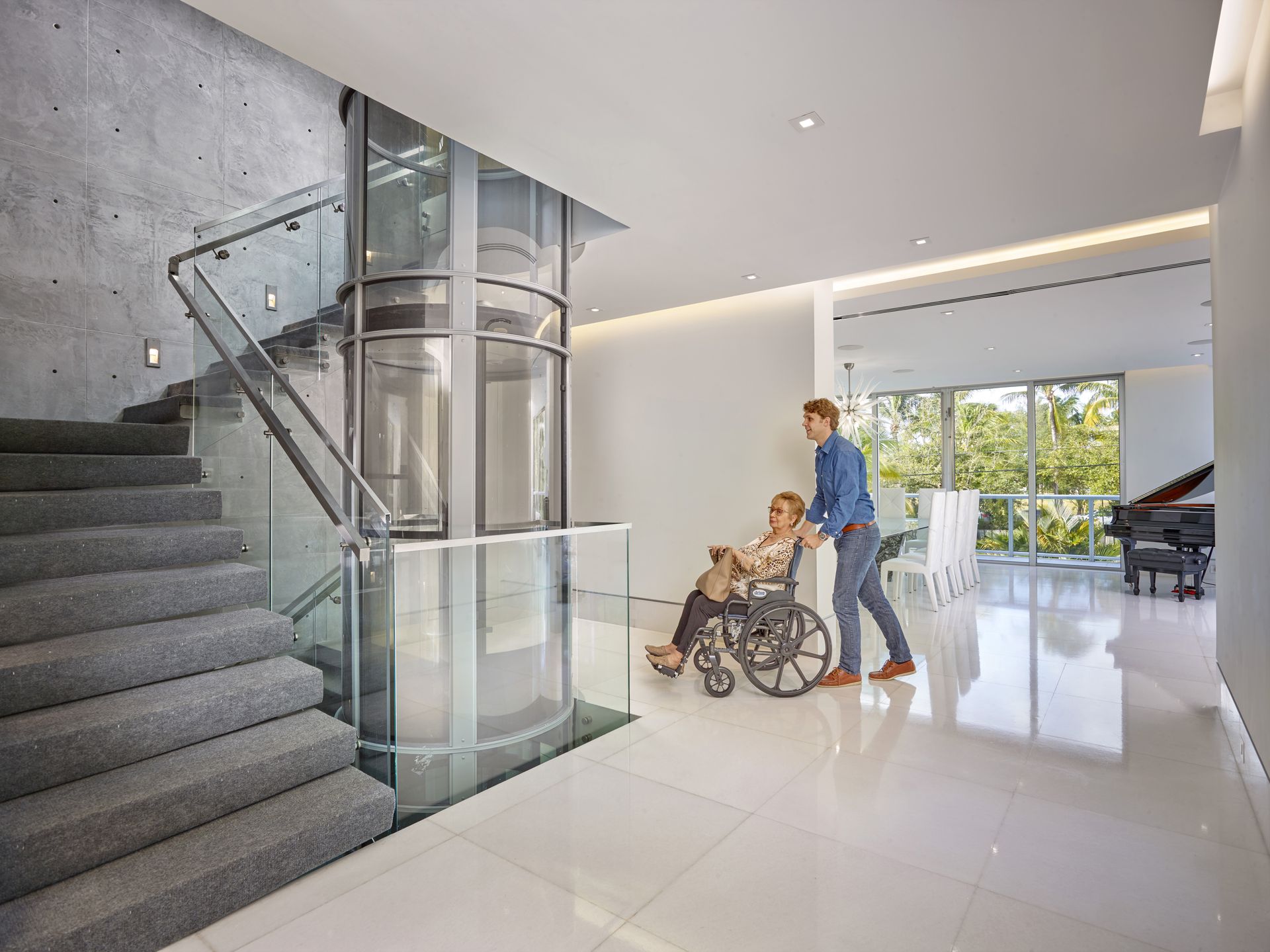 A person assisting someone in a wheelchair toward a glass residential elevator next to a staircase in a modern home.