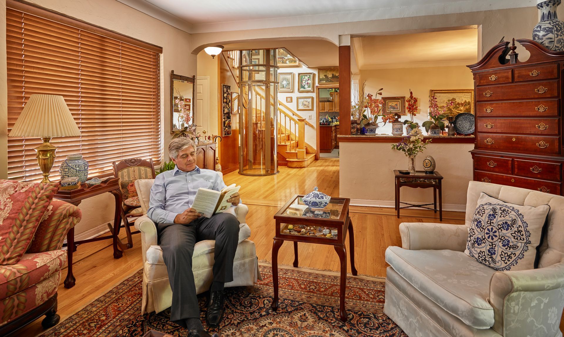 A person reads a book in a sunlit, traditional living room with hardwood floors, an area rug, and wooden furniture.