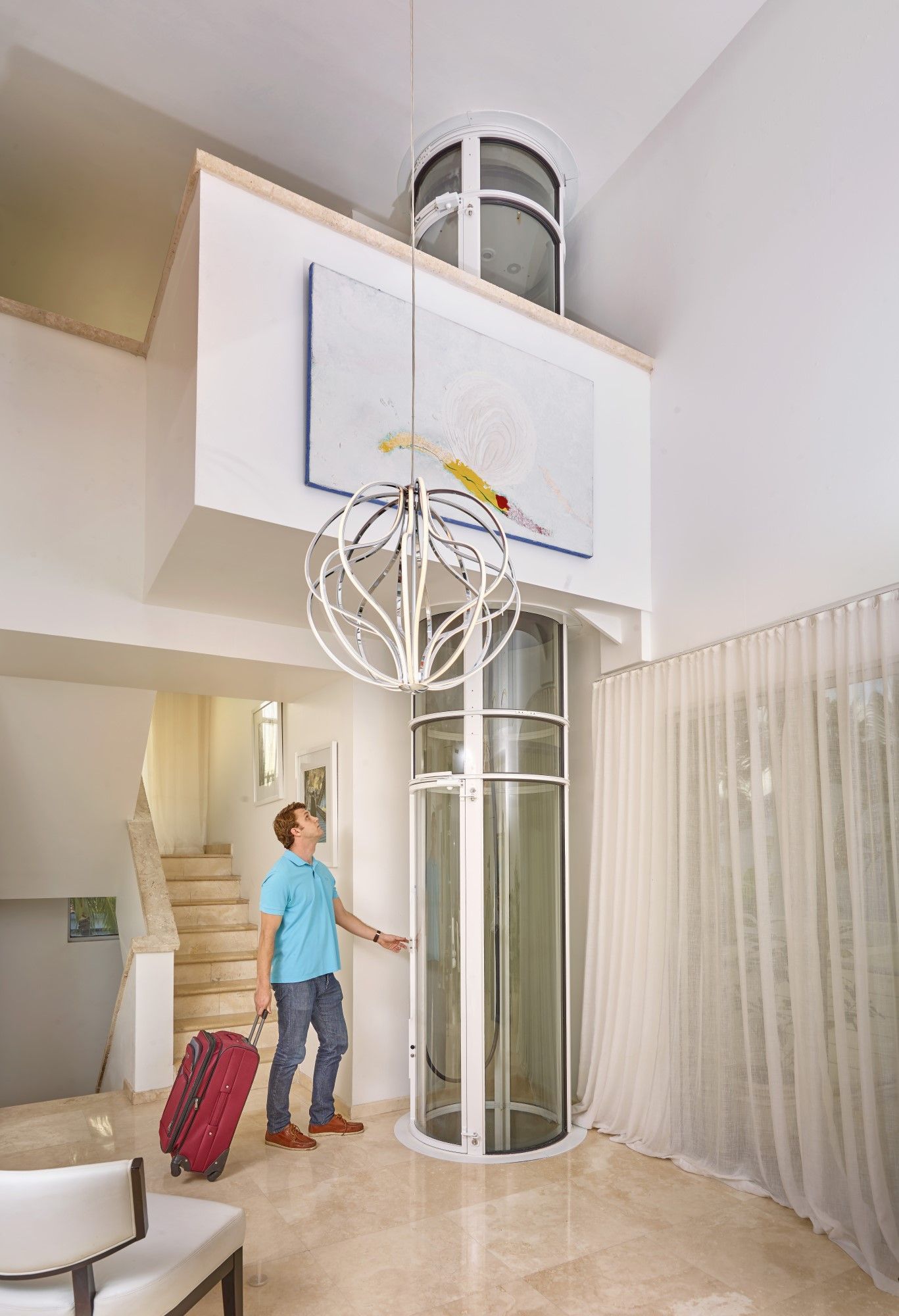 A person with a rolling suitcase stands in a bright, modern living room next to a cylindrical, glass residential elevator.