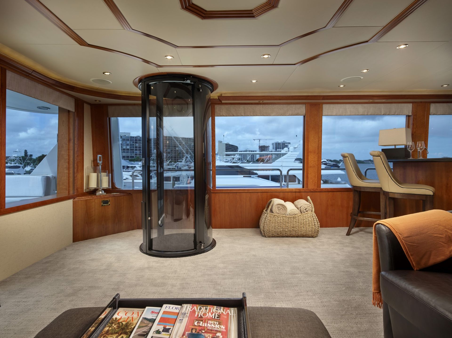 A spacious, modern yacht interior with a glass elevator, wood-paneled walls, carpeted floors, and marina views.