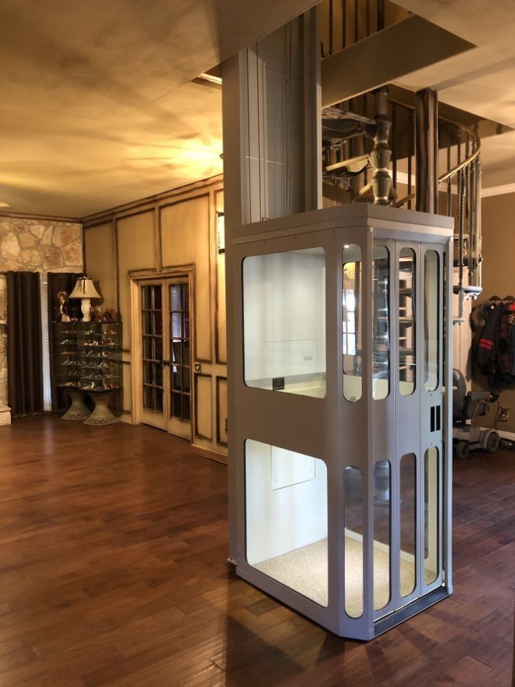 A residential elevator with white framing and glass panels standing in a room with wood floors and a spiral staircase.