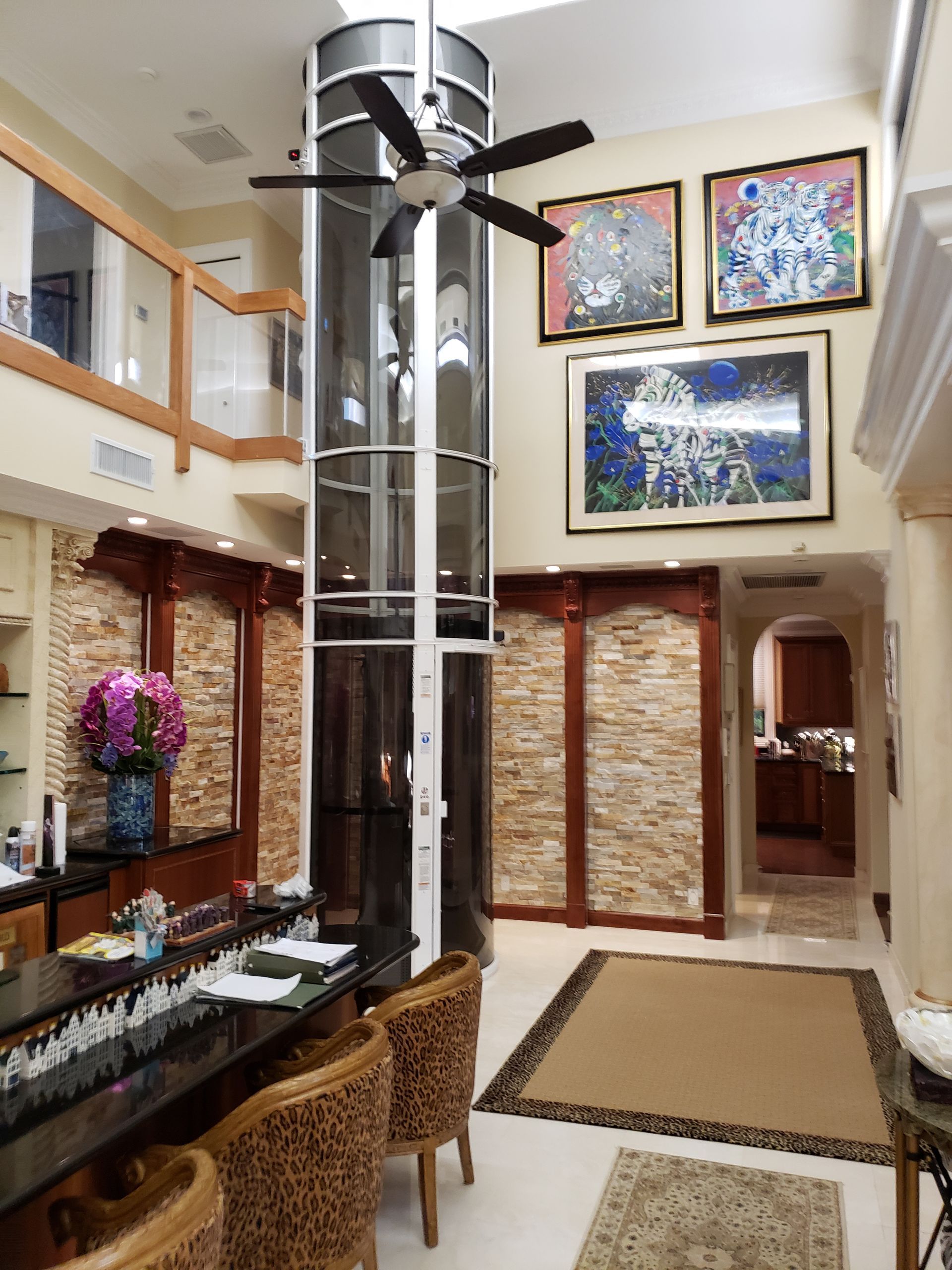 A glass cylindrical elevator stands in a room with stone walls, wooden accents, leopard-print chairs, and artwork.