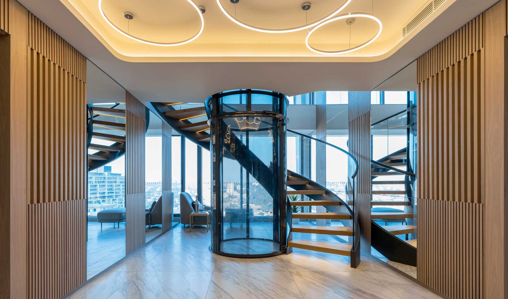 A modern interior features a central glass elevator surrounded by a curved, floating staircase with a wood-paneled lobby.