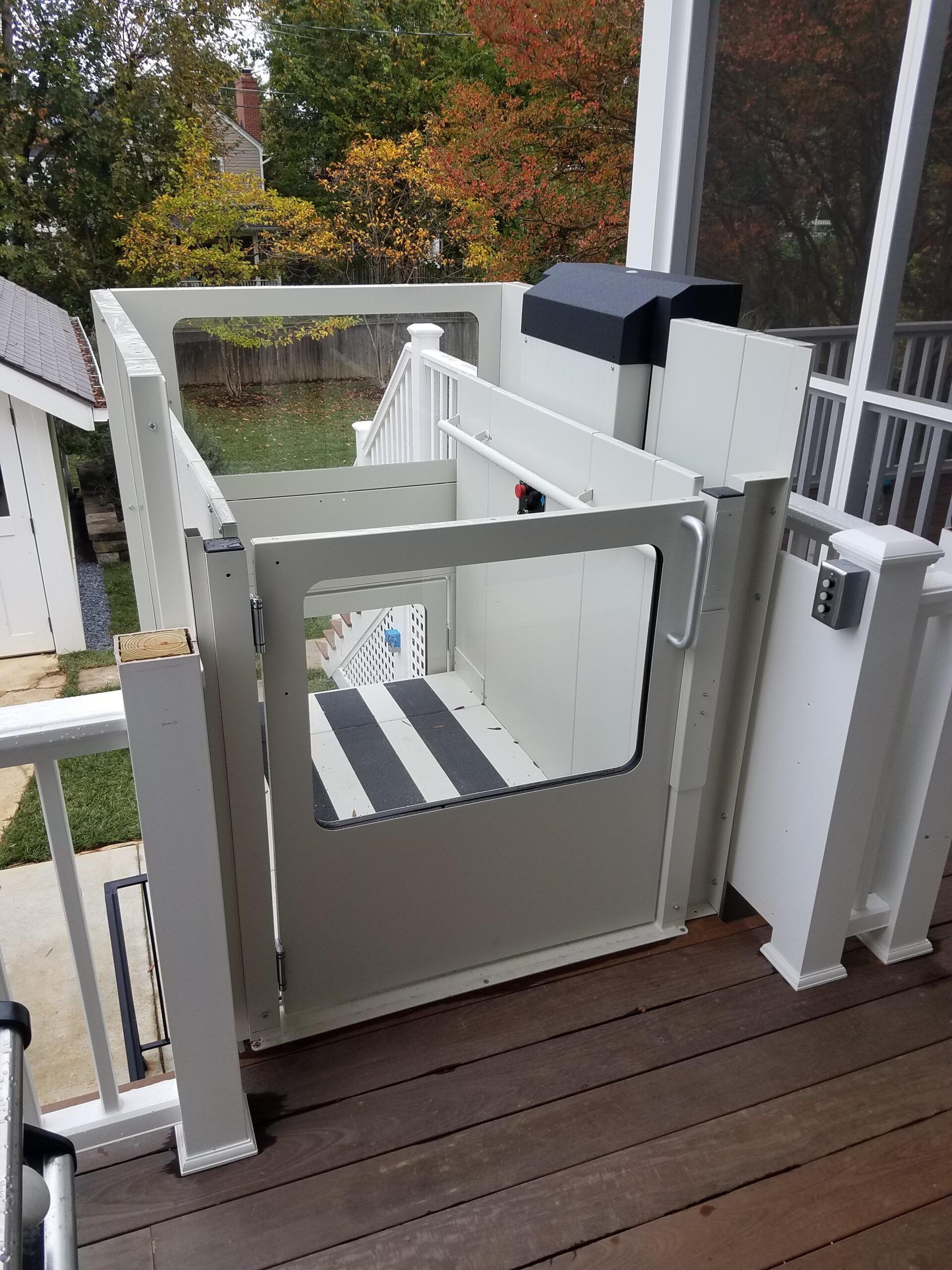 A tan outdoor wheelchair lift installed on a wooden deck, featuring a gated platform and a black motor cover.