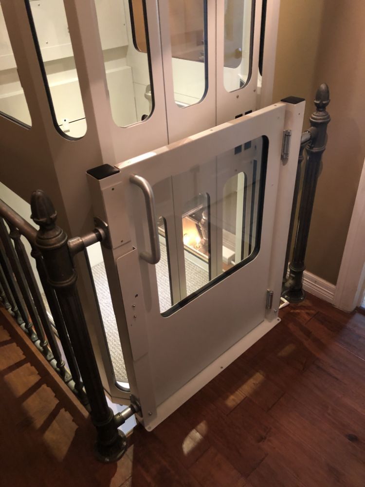 A residential elevator behind a white framed glass gate, positioned at a wooden staircase landing.