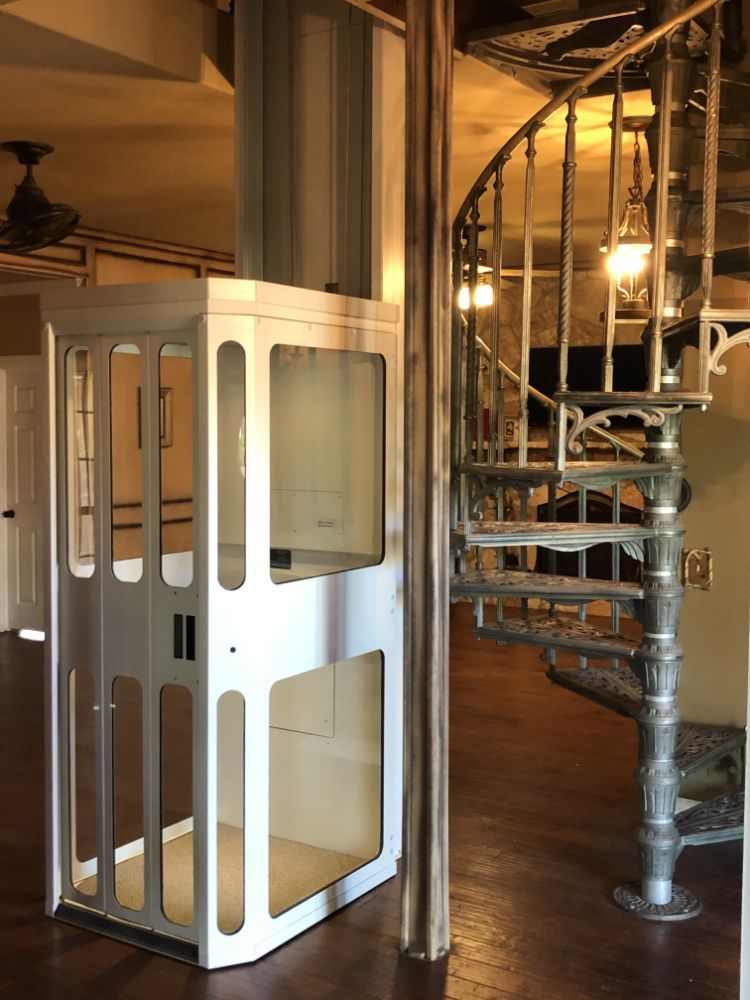A white, glass-walled residential elevator stands next to a decorative metal spiral staircase in an indoor room.