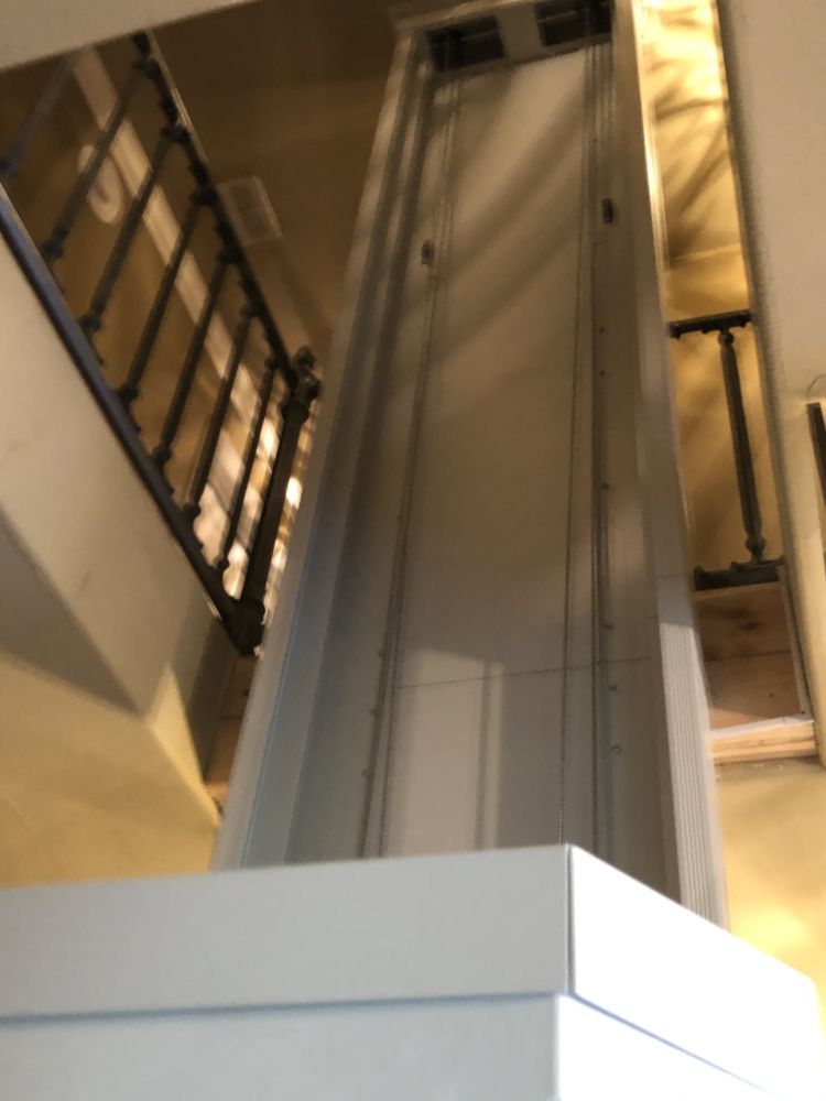 A view looking down into the open, vertical shaft of a residential elevator or lift, set within a staircase area.