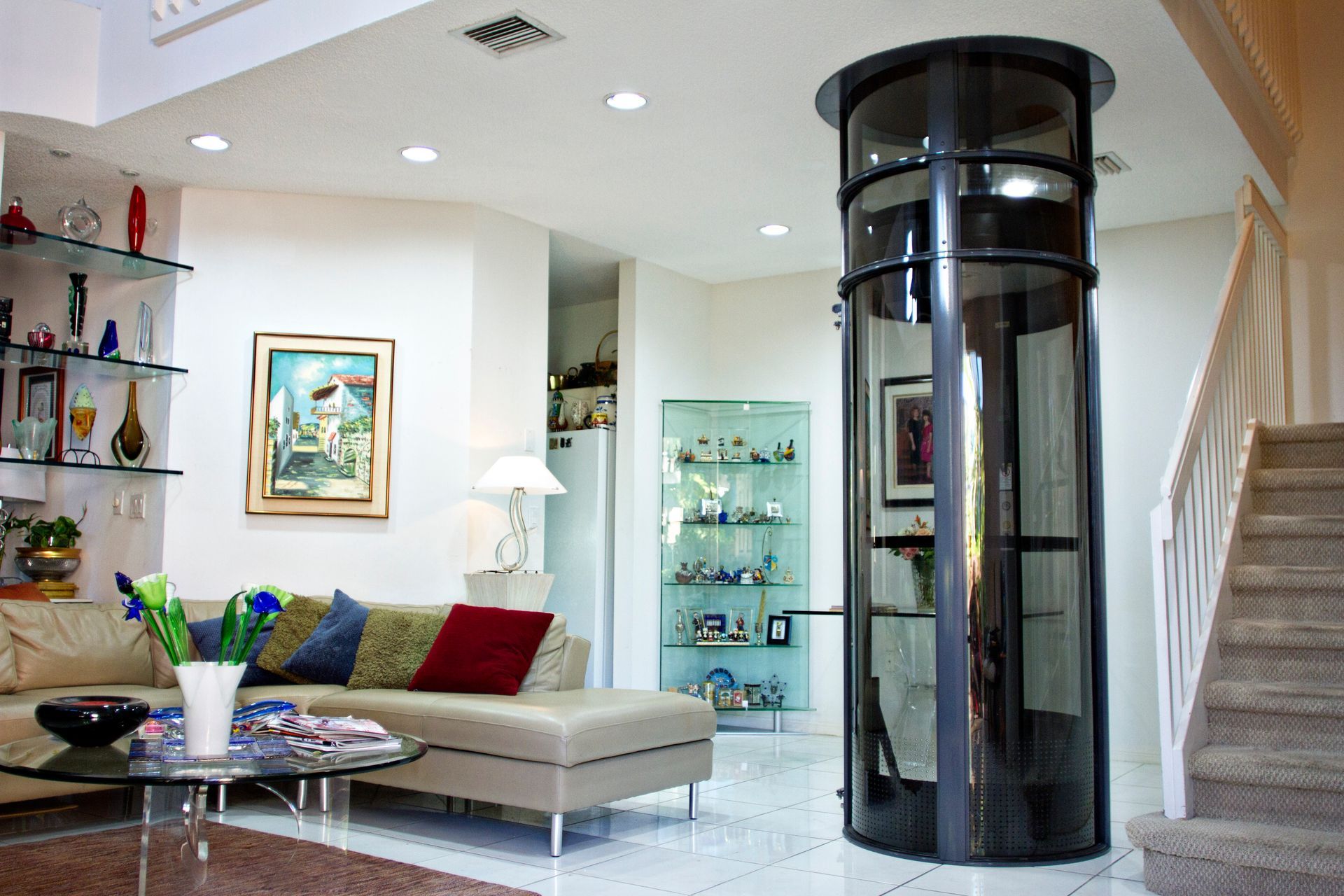 A modern, cylindrical glass home elevator stands in a living room next to a staircase and a sofa with colorful pillows.