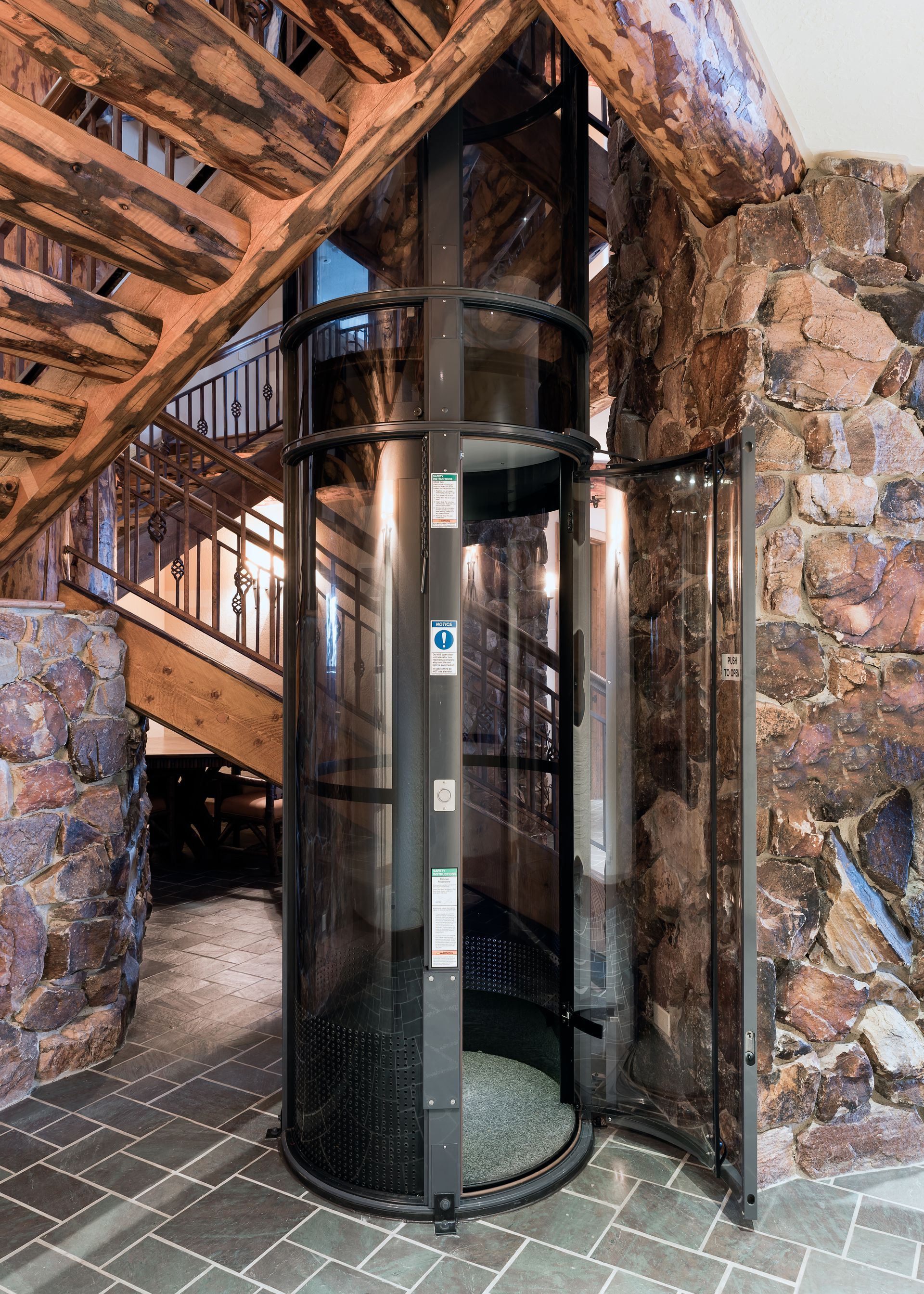 A cylindrical glass residential elevator positioned between a rustic stone wall and a wooden staircase.