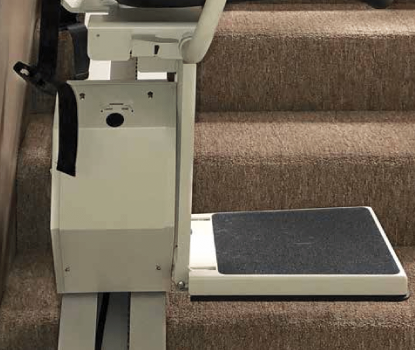 A brown and white stairlift mounted on a curved rail in a home with a wooden floor and a green potted plant nearby.